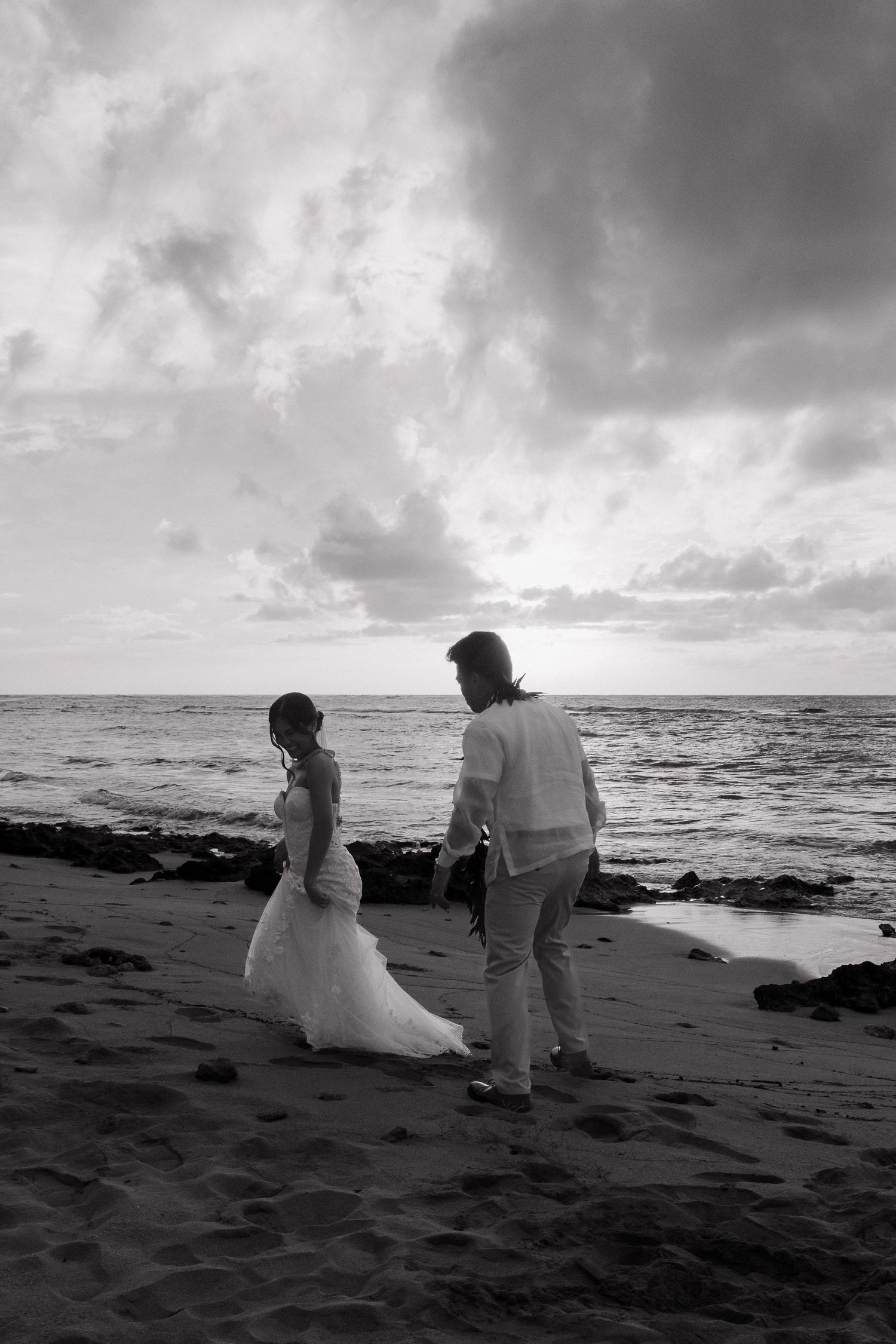 Black and white photo of a bride and groom walking hand in hand on a sandy beach at sunset.