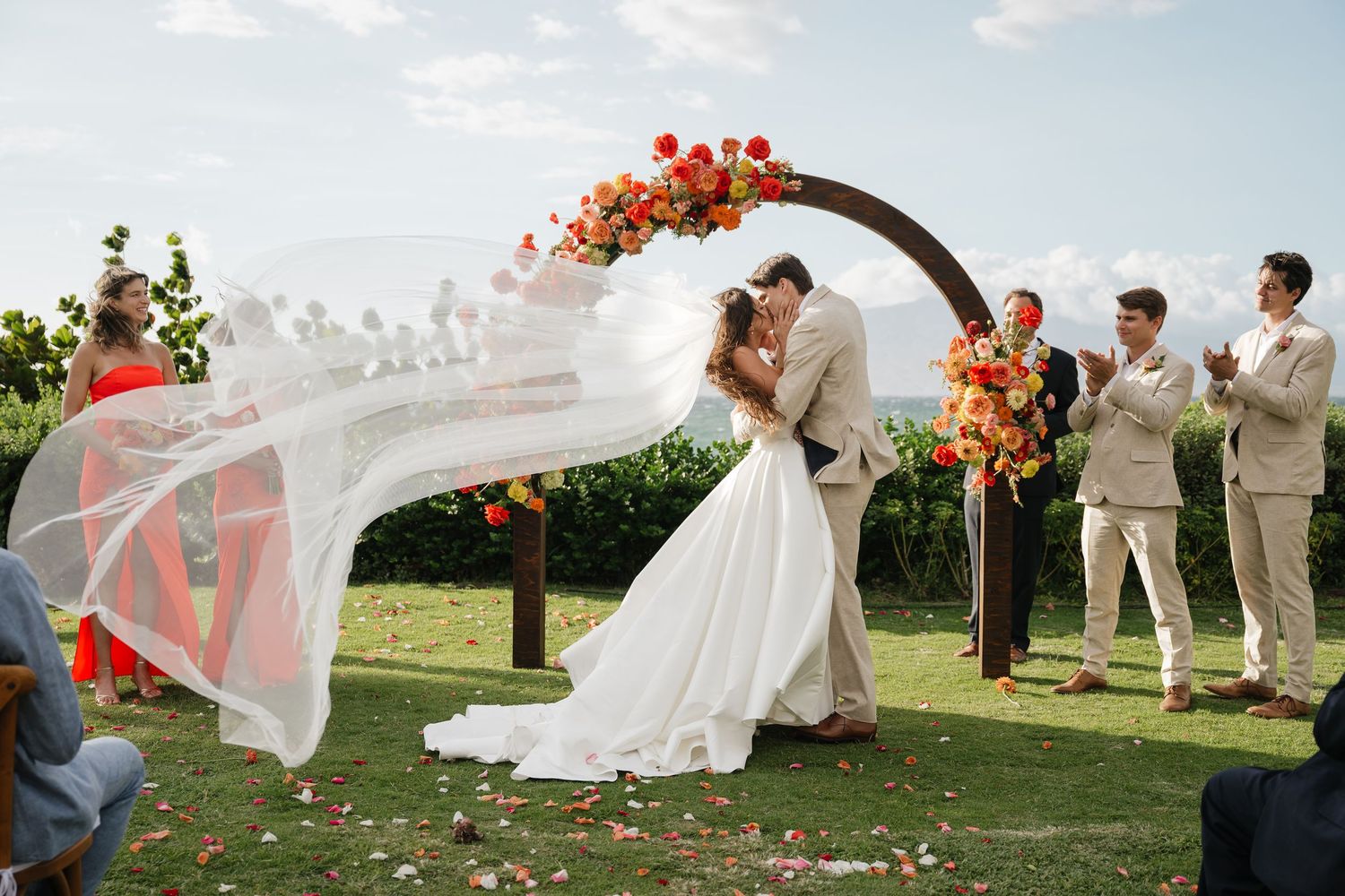 A wedding party poses together outdoors as wind catches a flowing veil and dress in the sunlight.