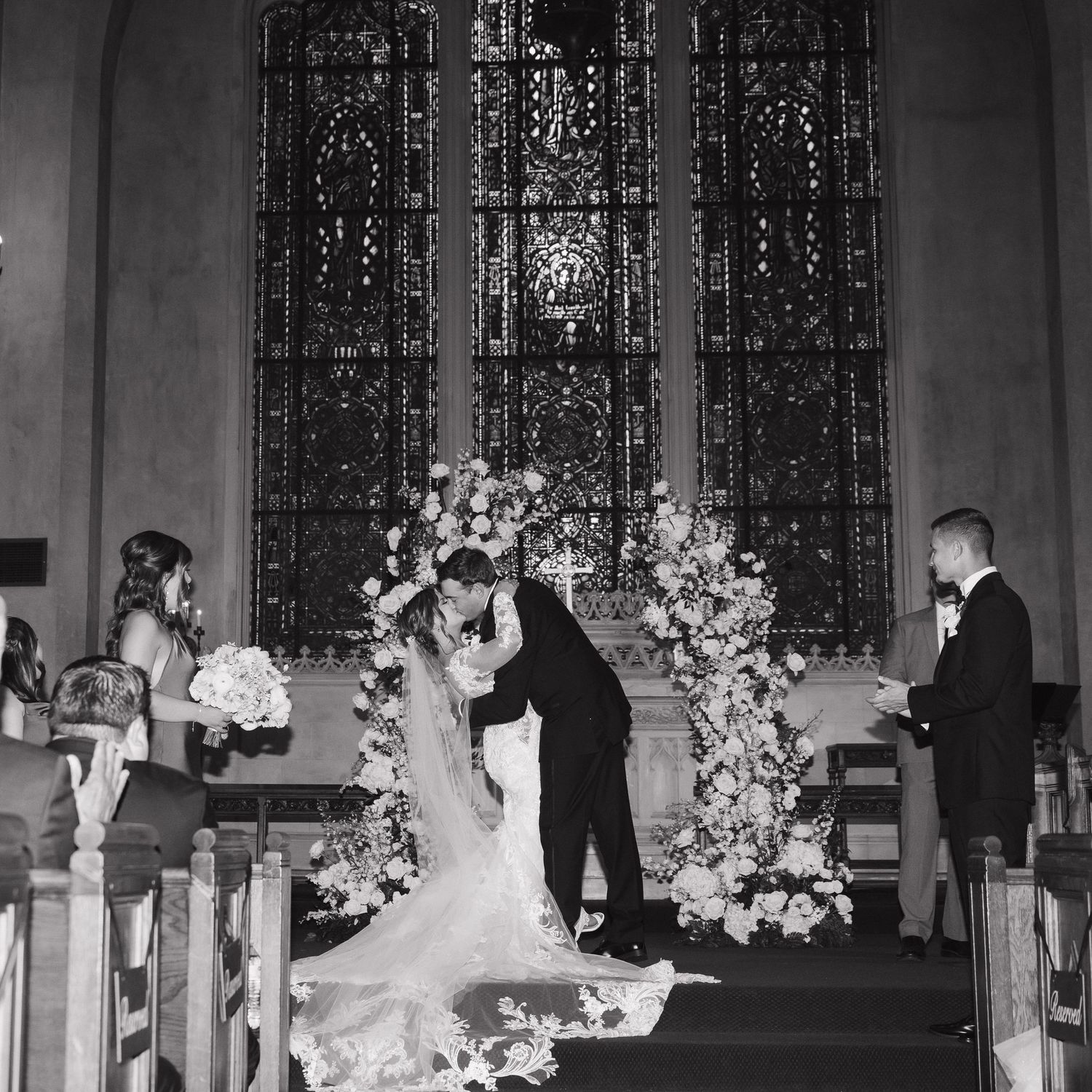 A wedding ceremony takes place in a church with stained glass windows and floral arrangements.