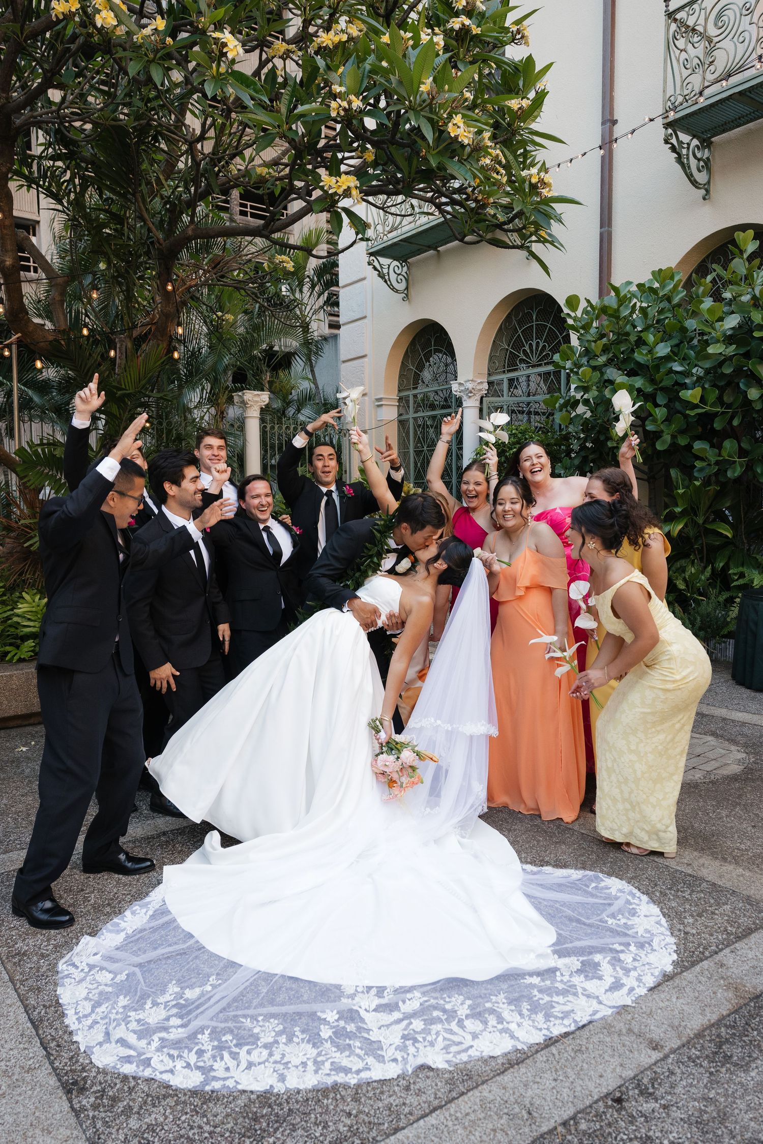 Bride and groom share a dip kiss surrounded by cheering wedding party in colorful attire outside elegant venue.