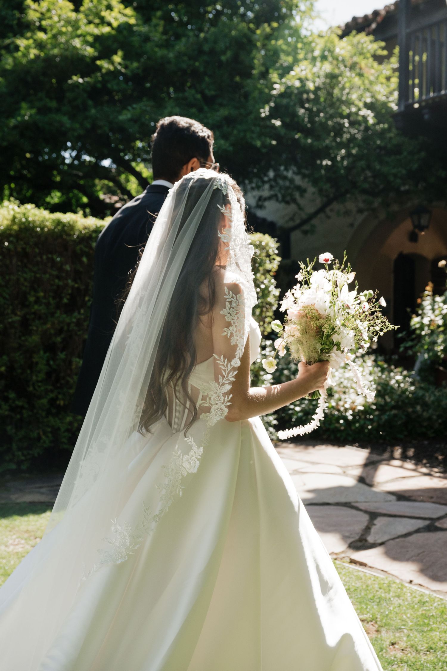 Bride in lace wedding gown and veil holding white bouquet walks with groom in garden setting.