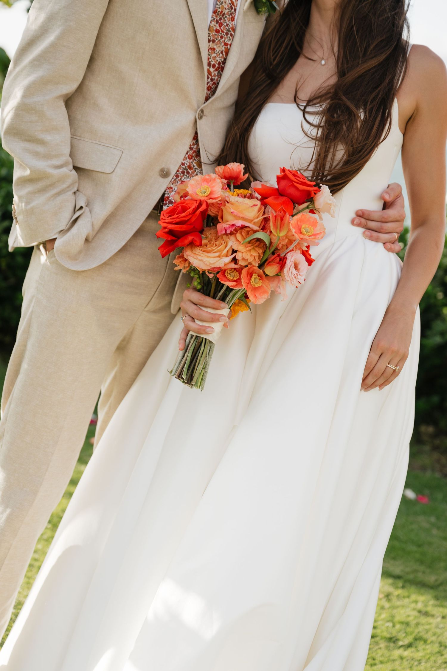 Wedding couple in tan suit and white dress hold coral bouquet in garden setting.