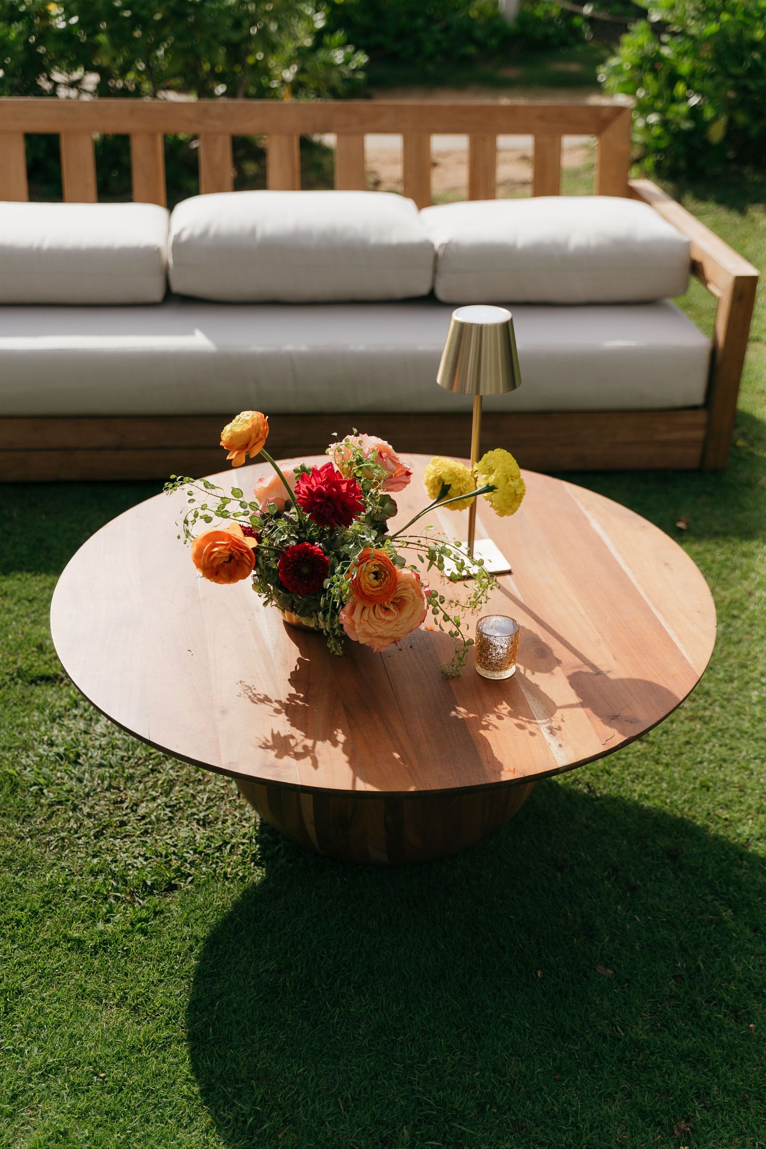 Outdoor patio setting with round copper table, floral arrangement and modern lamp next to wooden daybed with beige cushions.
