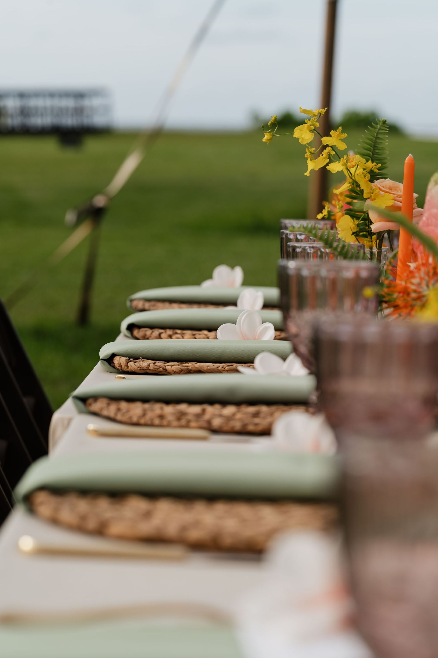 Elegant outdoor wedding table setting with sage green napkins, woven placemats, pink glassware, and yellow floral centerpieces.