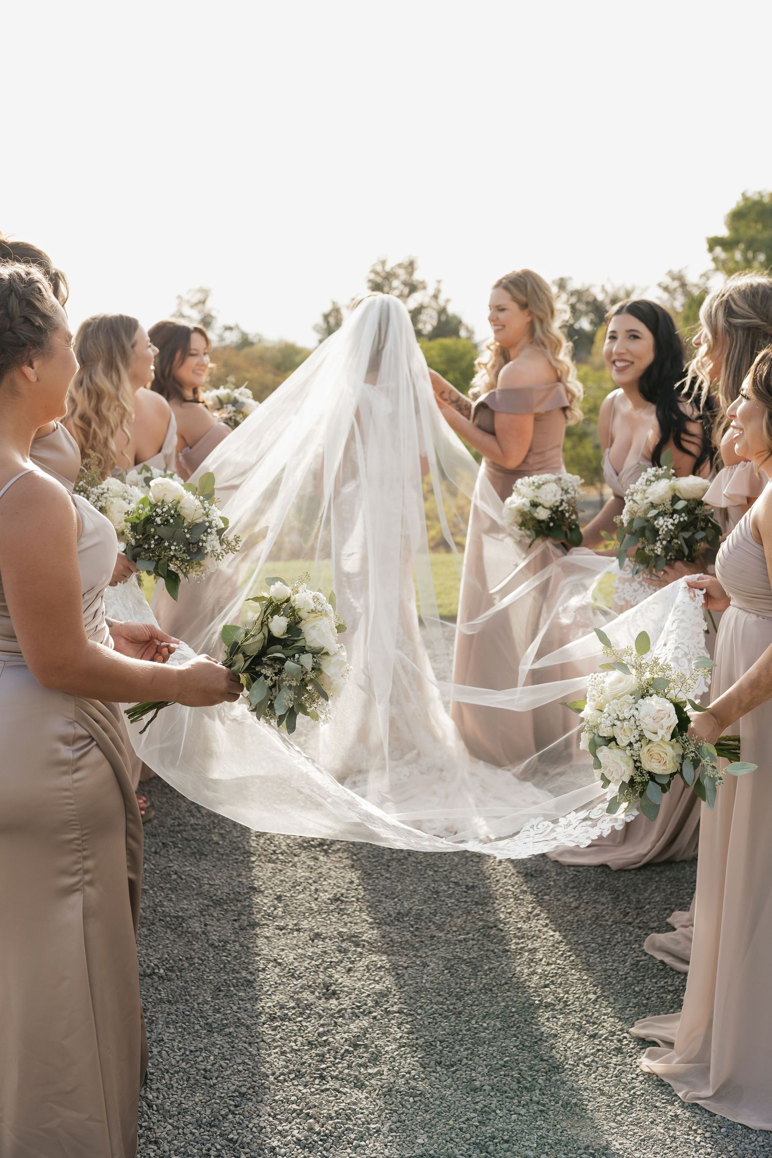 Bride in flowing lace gown walks with bridesmaids in neutral dresses along gravel path.