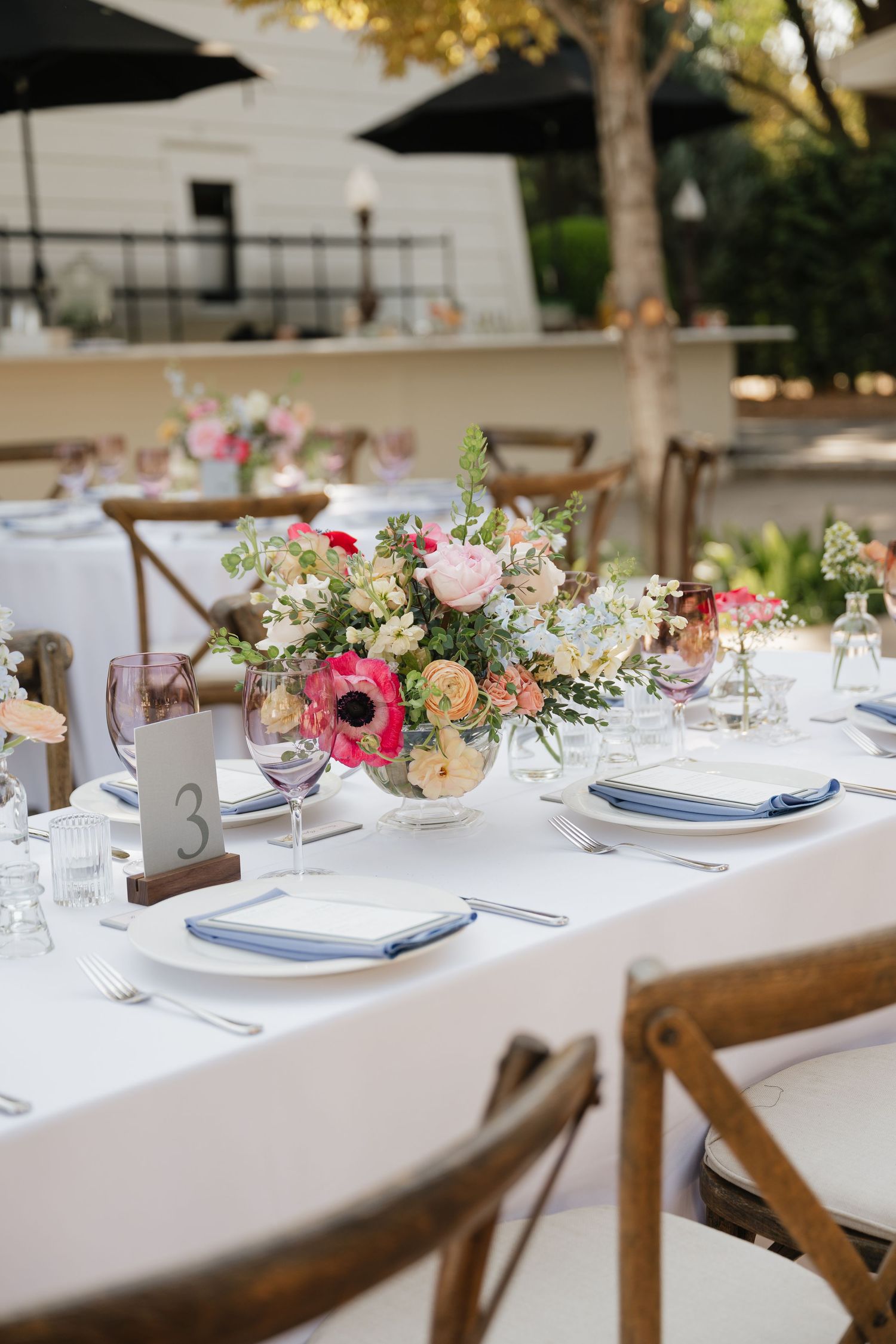 Long dining table set with white linens and colorful floral centerpieces for an outdoor wedding reception.