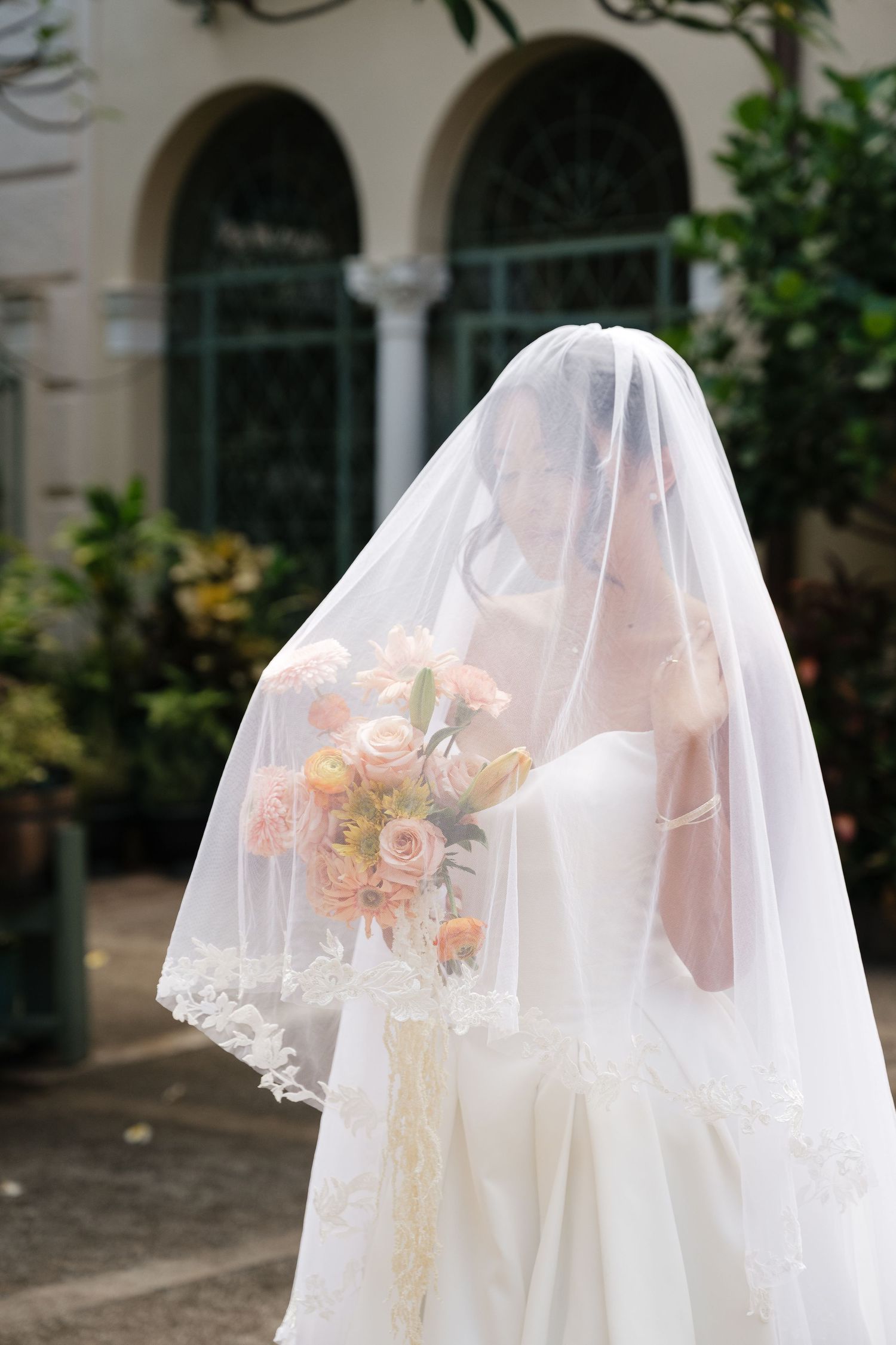 Bride in white wedding dress and lace veil holding pink rose bouquet outdoors near elegant building.