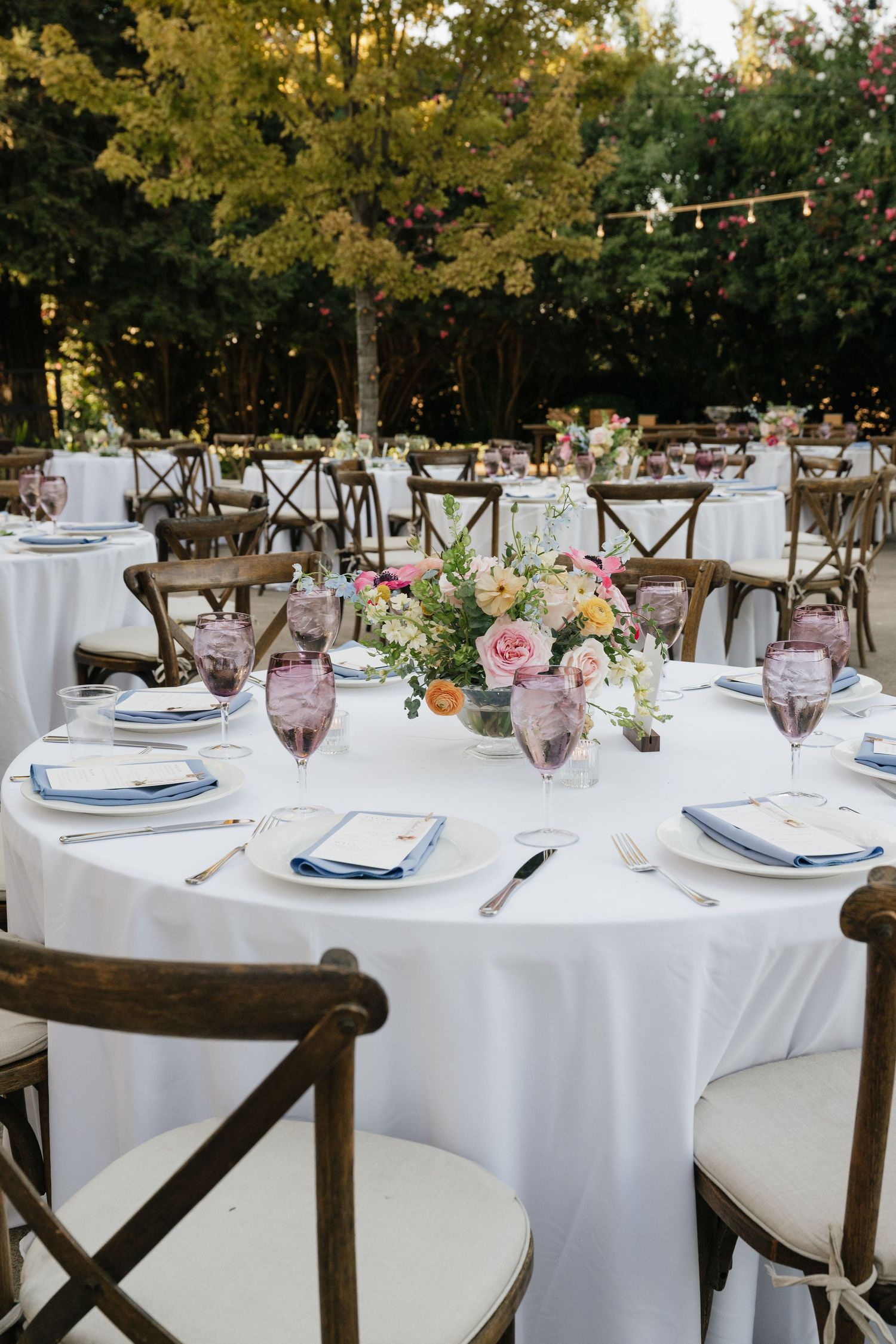 Elegant outdoor wedding reception setup with white tablecloths and wooden cross-back chairs.