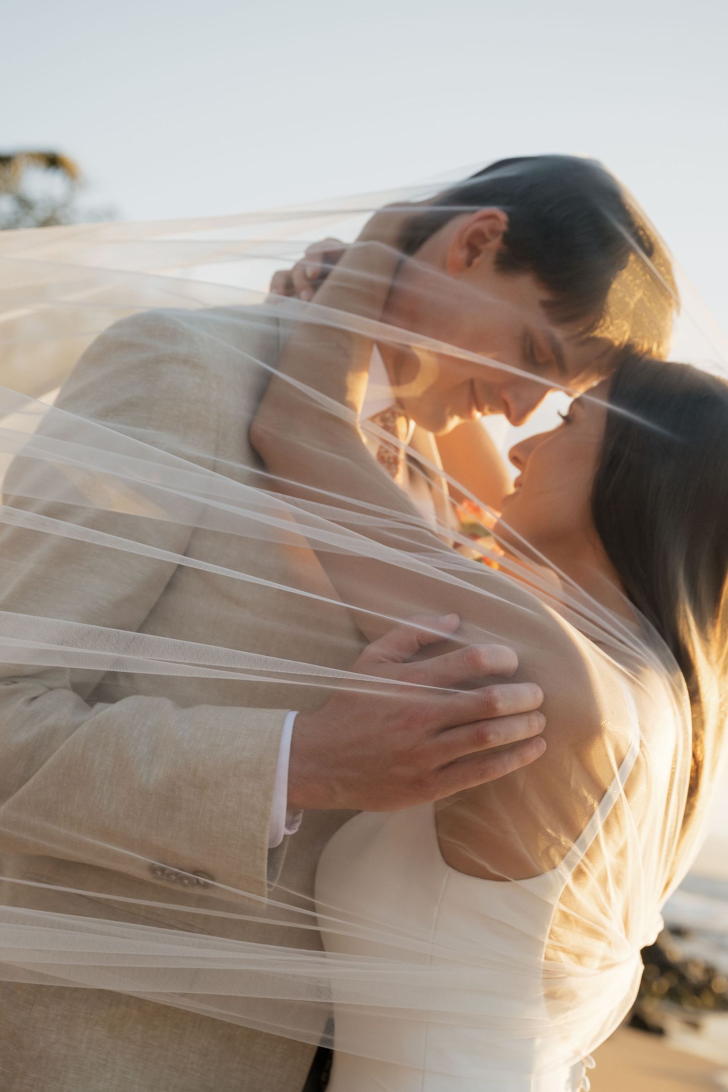 A romantic couple shares a tender kiss under a flowing white veil during a sunset wedding photoshoot.