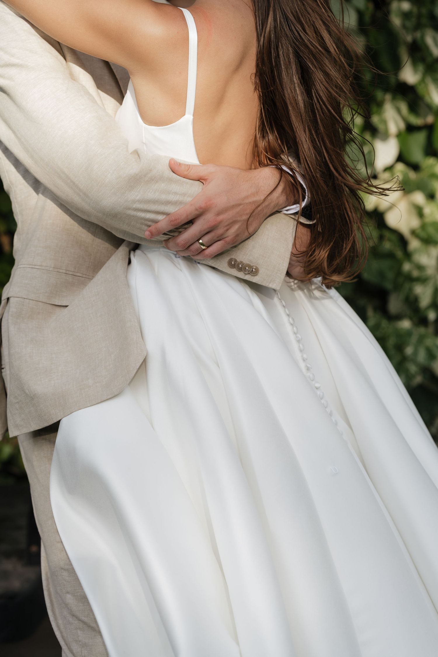 Close up details of a white wedding dress and light colored suit during an embrace.