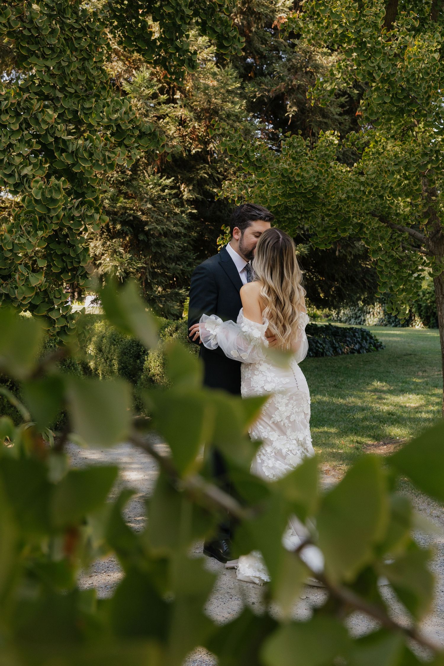 A romantic couple embraces in a lush garden setting with green foliage and natural sunlight filtering through the trees.