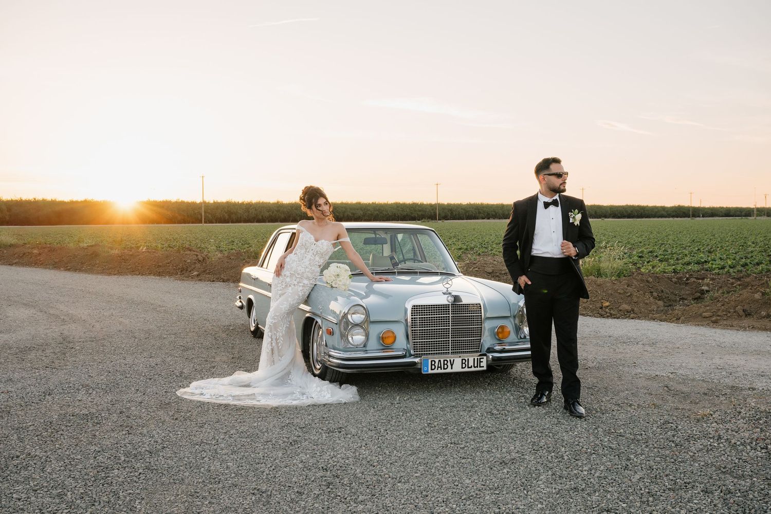 A wedding couple poses with a classic Mercedes-Benz car during sunset in an open field.