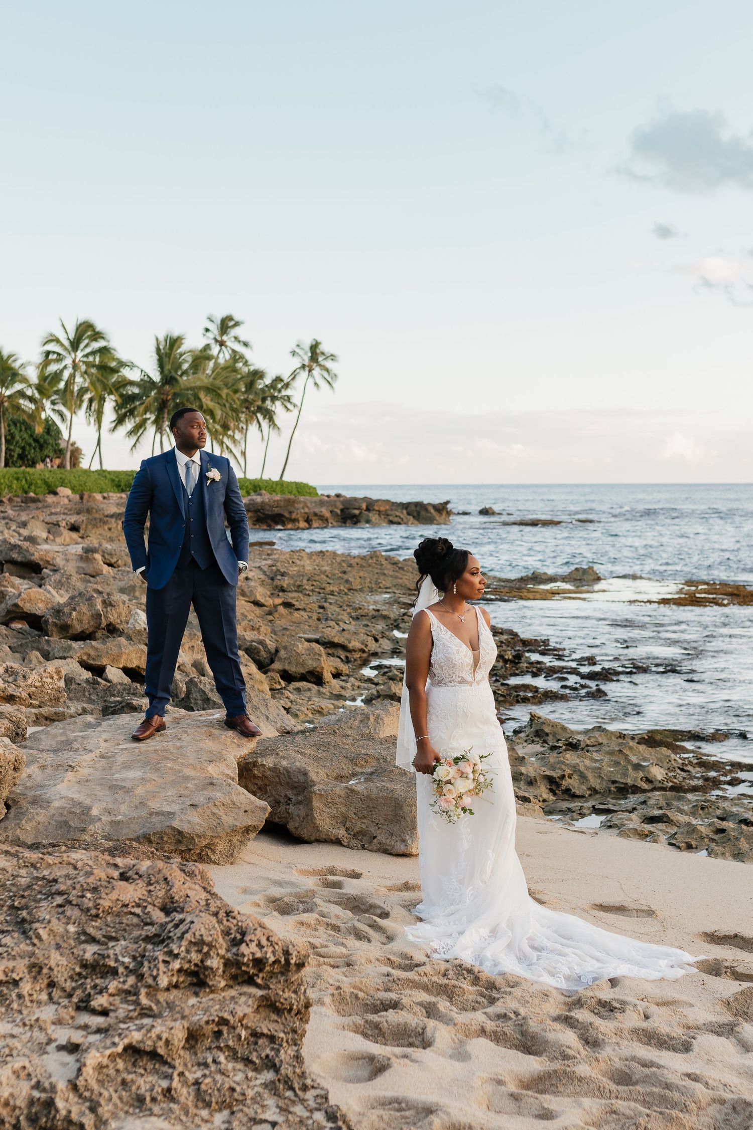 Couple poses for romantic wedding photos on a rocky beach with palm trees and ocean views.