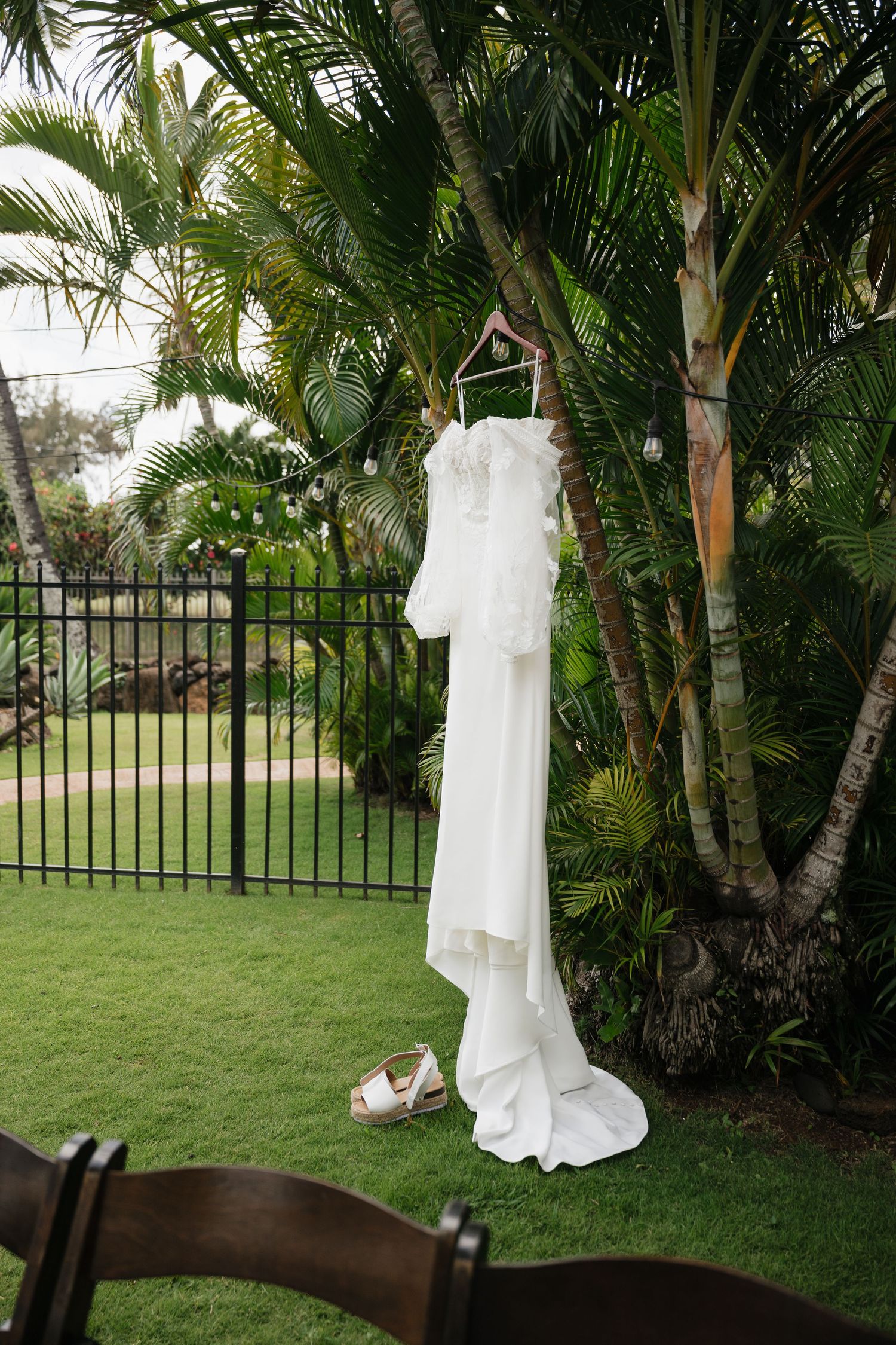 Wedding gown displayed on hanger among palm trees with iron fence in background.