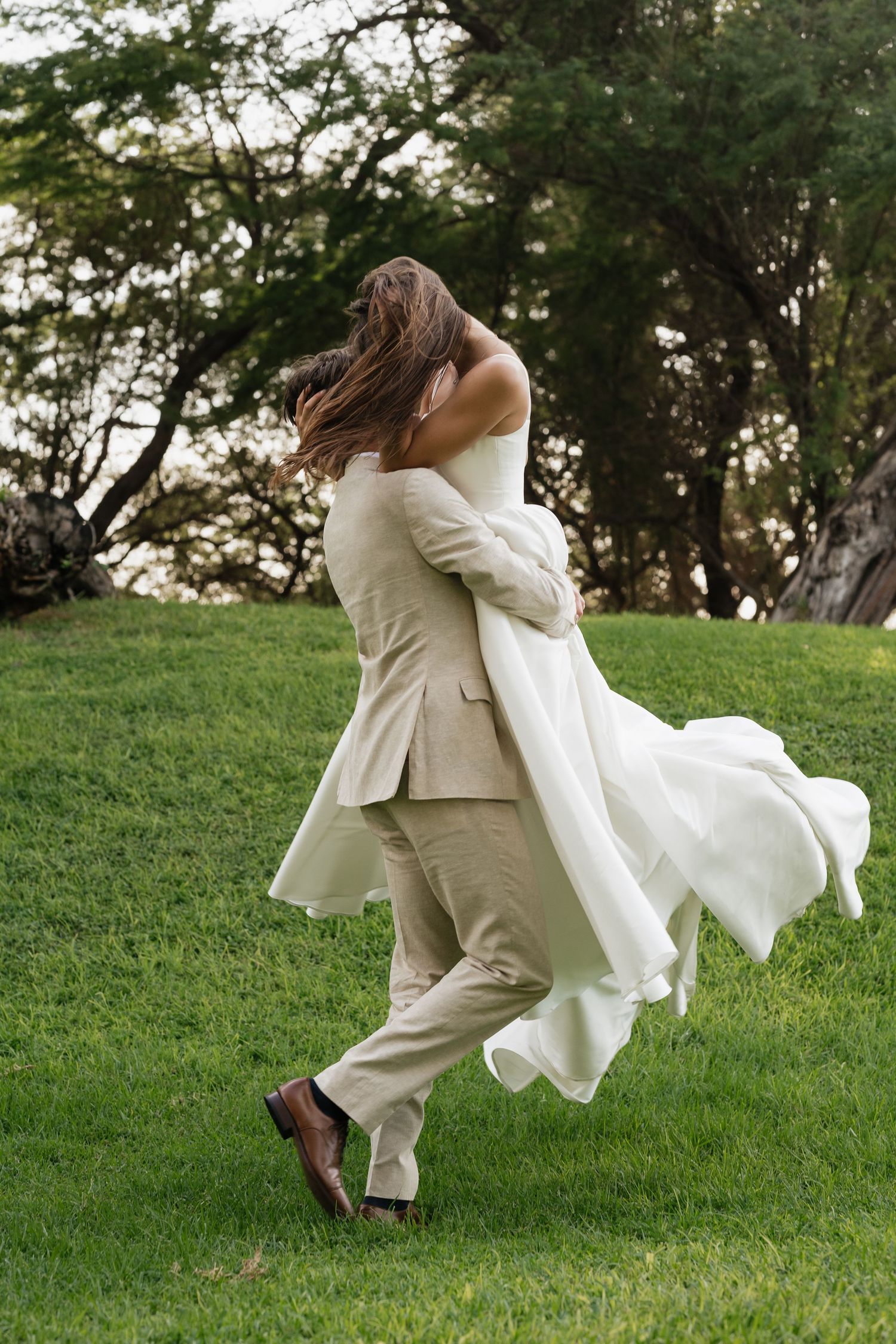 A romantic couple dances and spins playfully on a grassy hillside wearing coordinated cream-colored formal attire.