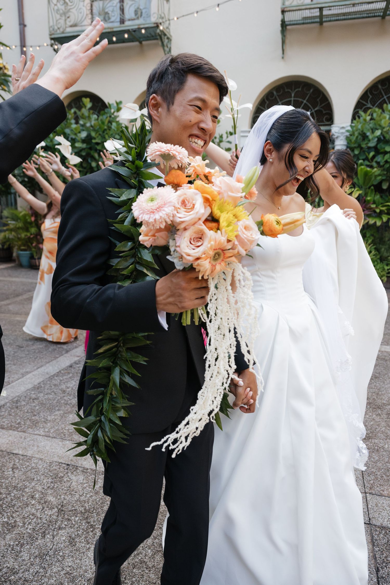 Smiling groom in black suit with lei holds peach bouquet beside bride in white gown after Hawaiian wedding ceremony.
