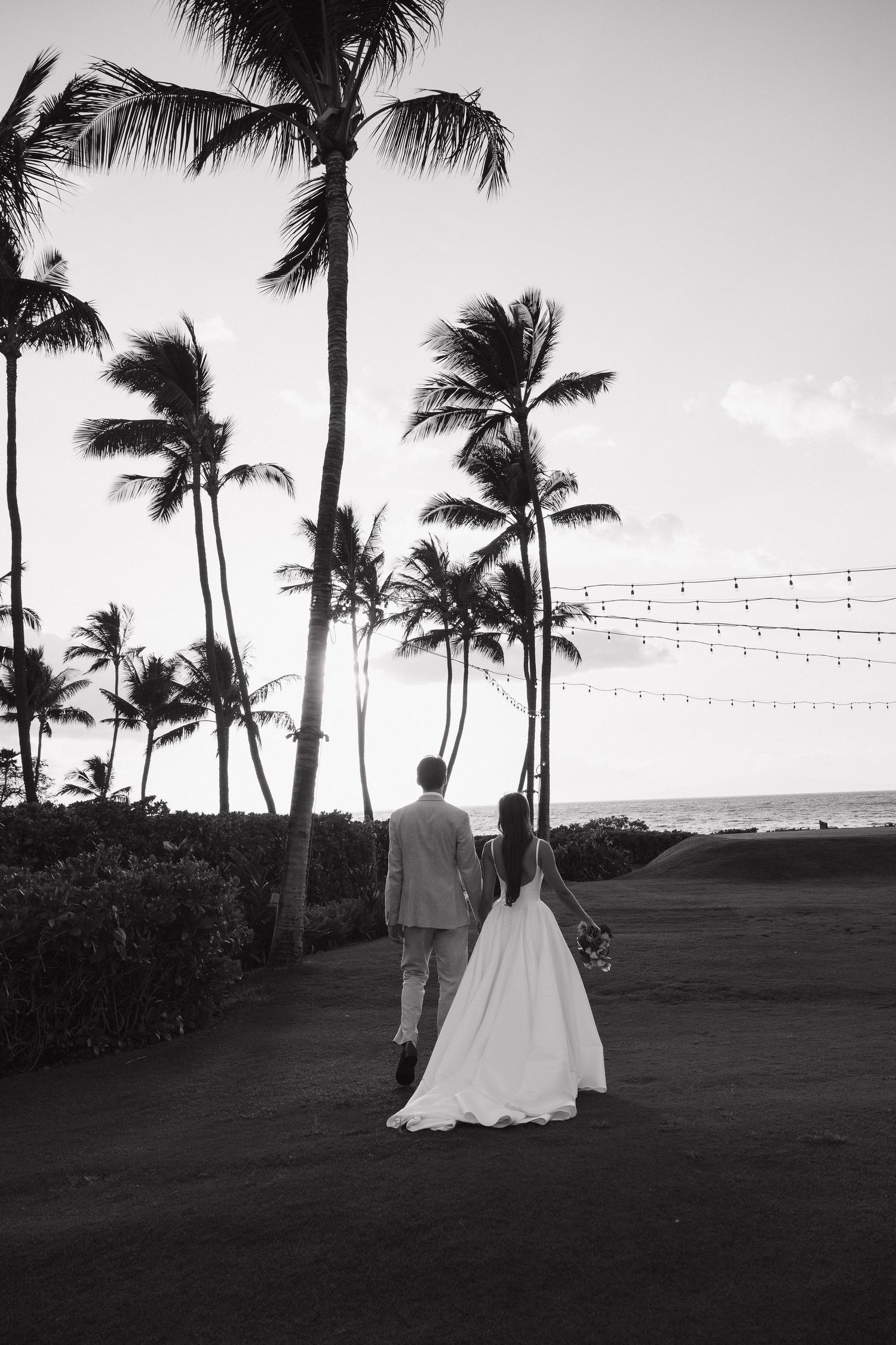 Black and white photo of a couple walking along a palm-lined beach path at sunset.