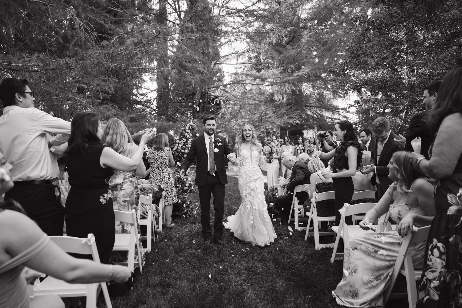 Wedding guests dance and celebrate outdoors during a festive reception in a wooded garden setting.