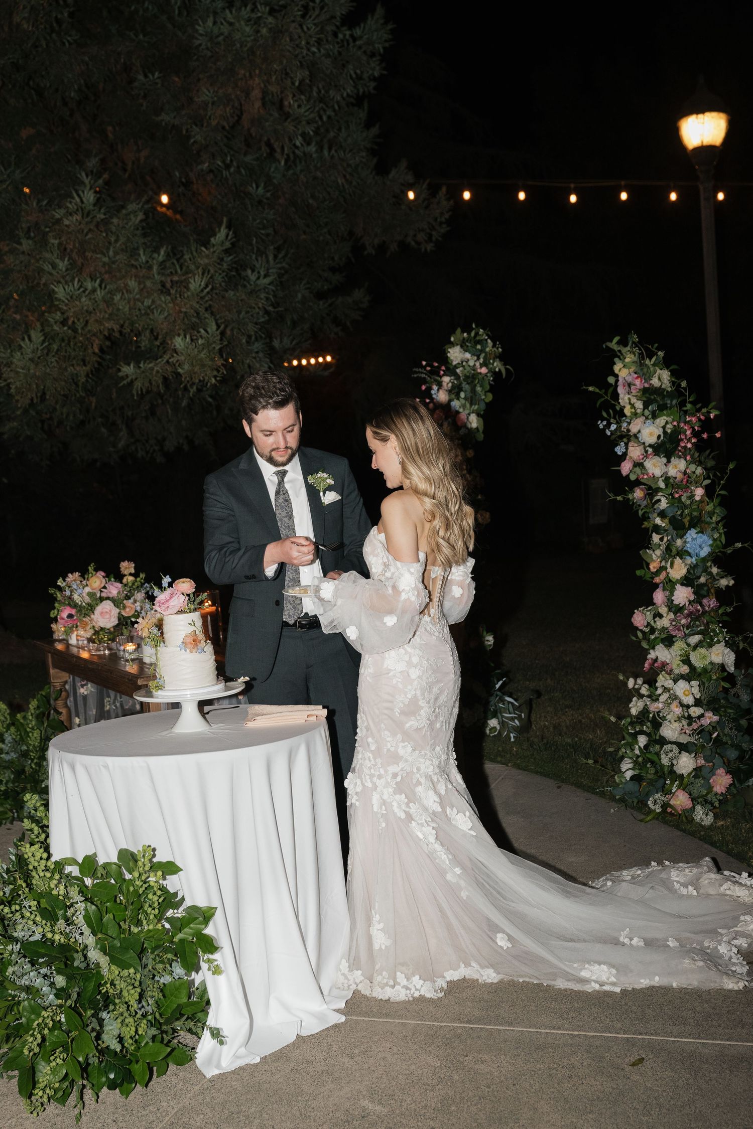 Couple shares a sweet moment while cutting their wedding cake outdoors at night surrounded by floral arrangements.
