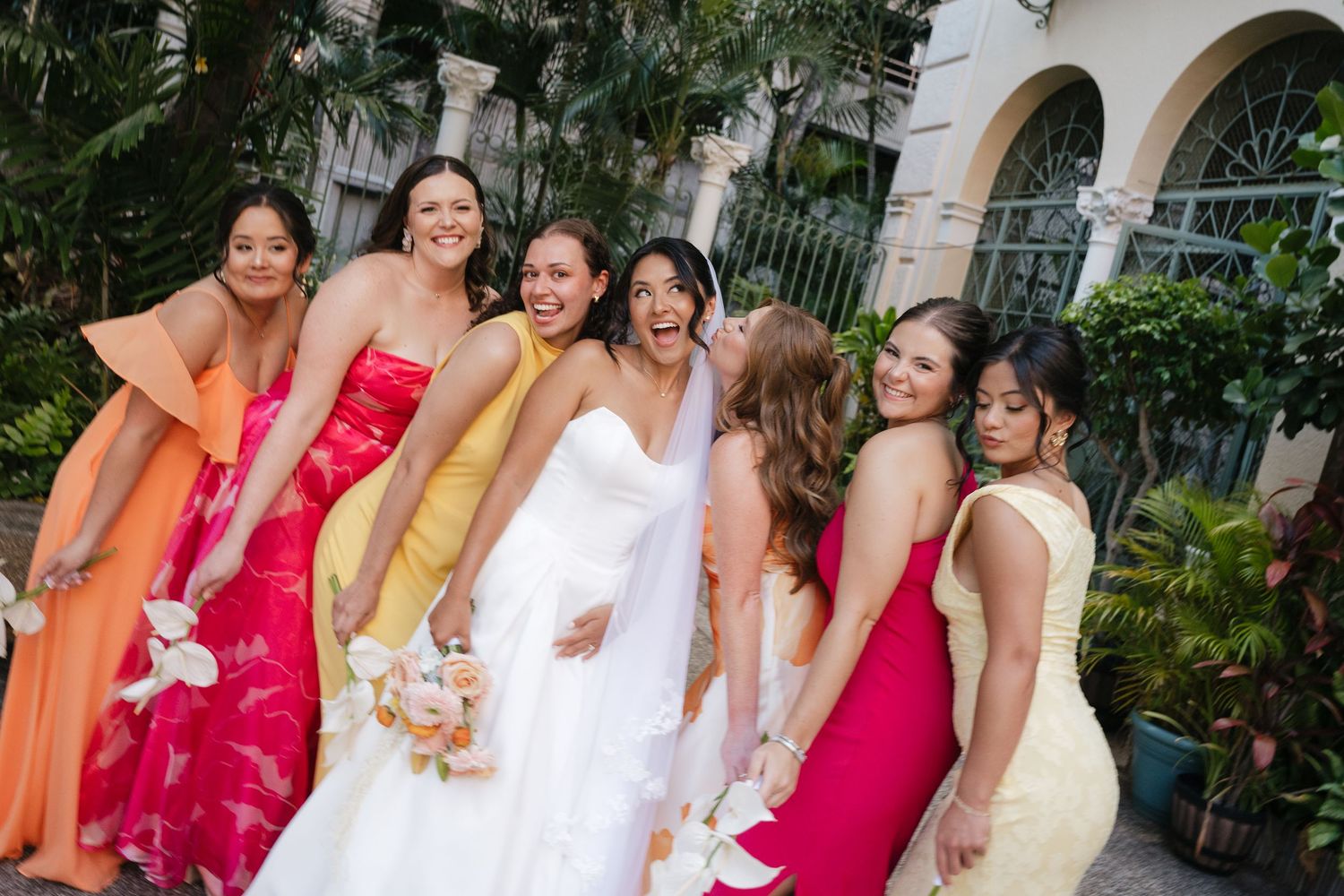 Bride in white gown poses joyfully with six bridesmaids in colorful dresses outdoors at a wedding celebration.