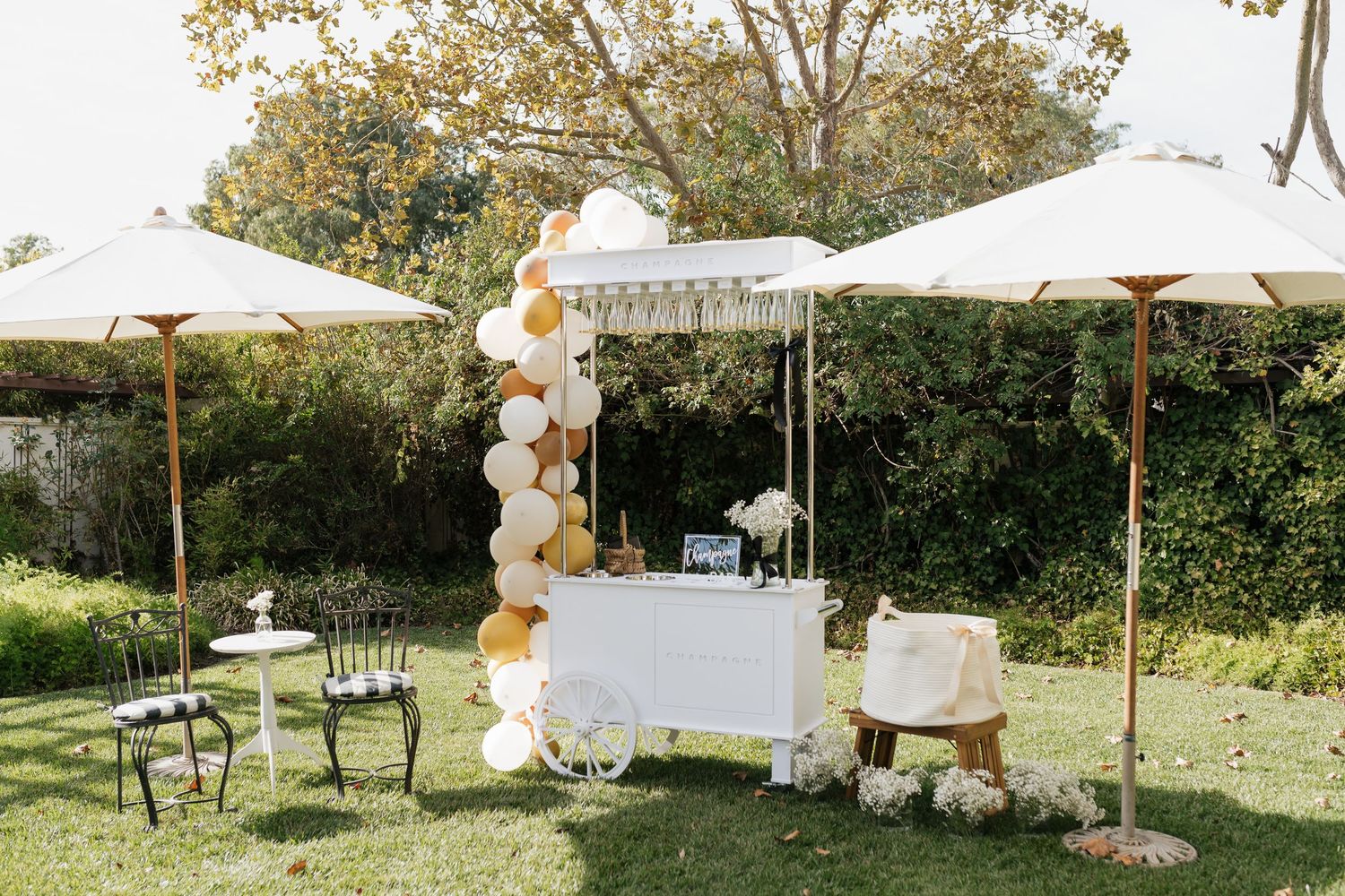 Outdoor wedding setup with white canopy, balloon arch and decorative display table under white umbrellas.