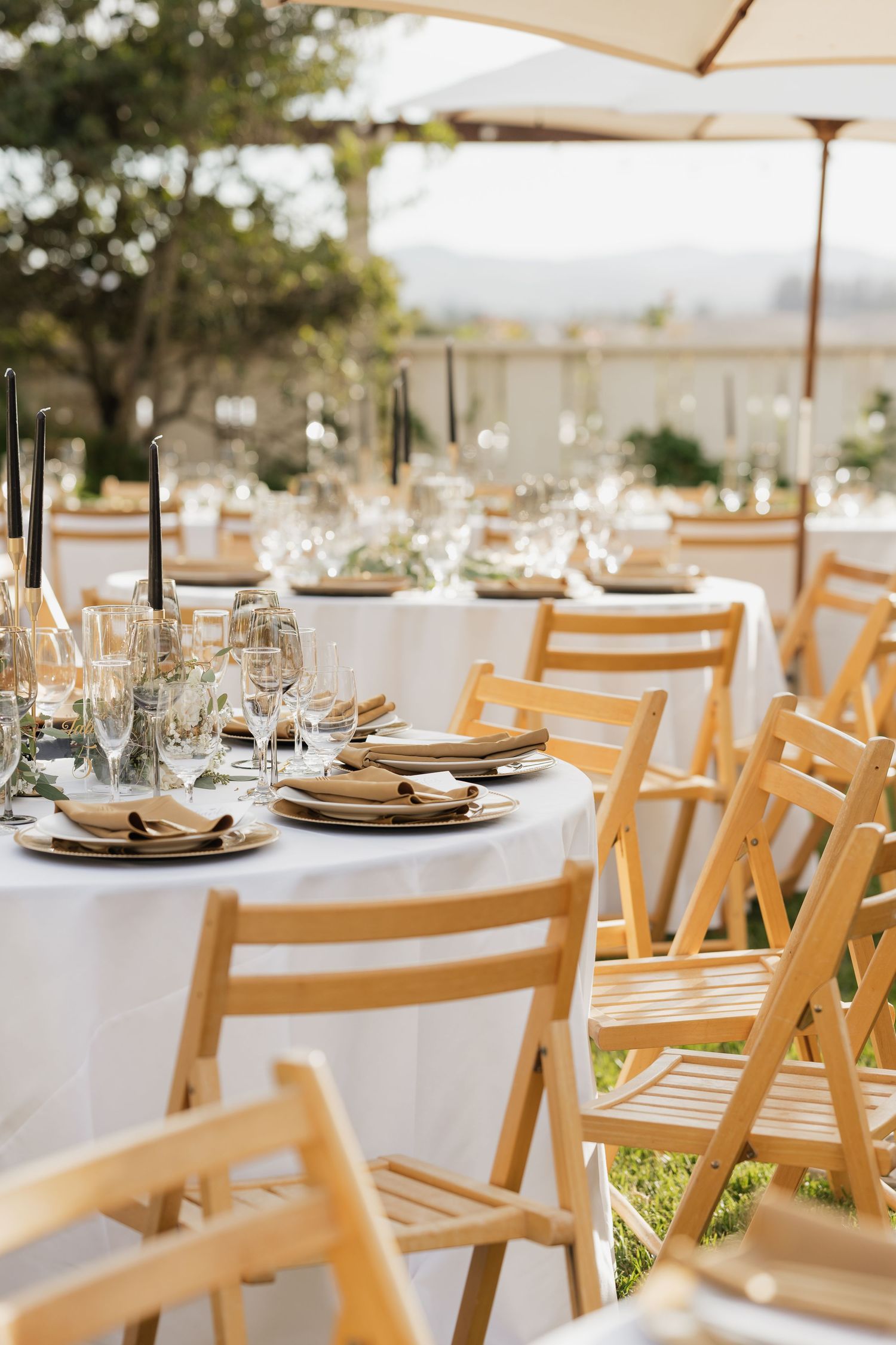 Outdoor reception tables set with white linens and wooden chairs under market umbrellas.