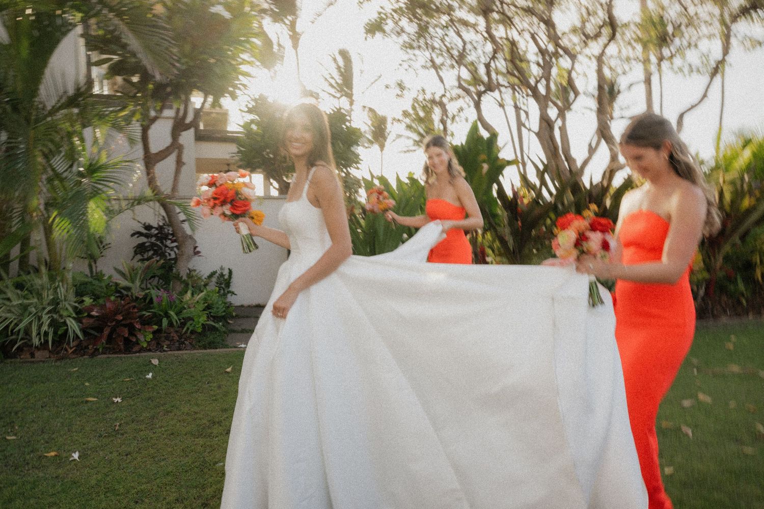 A wedding party in white dresses and bright coral gowns poses together in a lush garden setting.