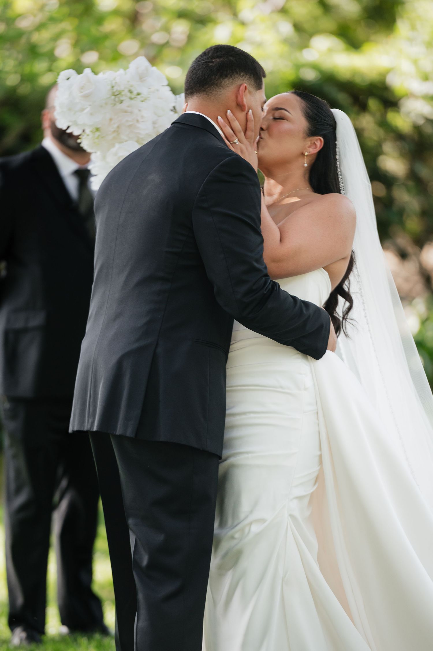 Bride and groom share their first kiss at outdoor wedding ceremony, bride in white gown with veil, groom in black suit.
