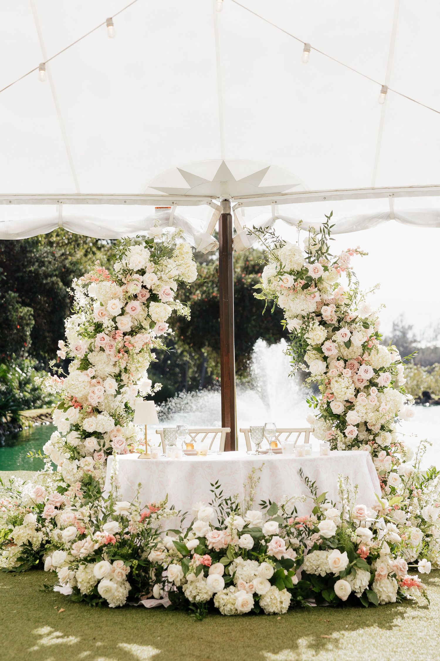 Elegant wedding sweetheart table adorned with lush white and pink floral arch and hydrangea ground arrangements under a tent.