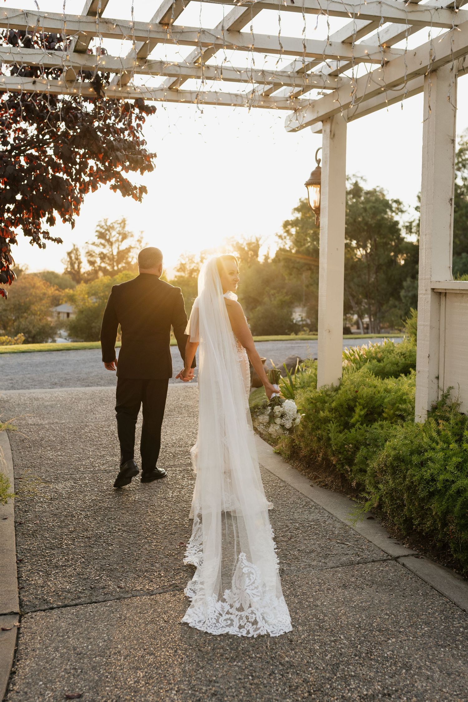 A wedding couple walks away from camera under a pergola at sunset with a trailing lace veil.