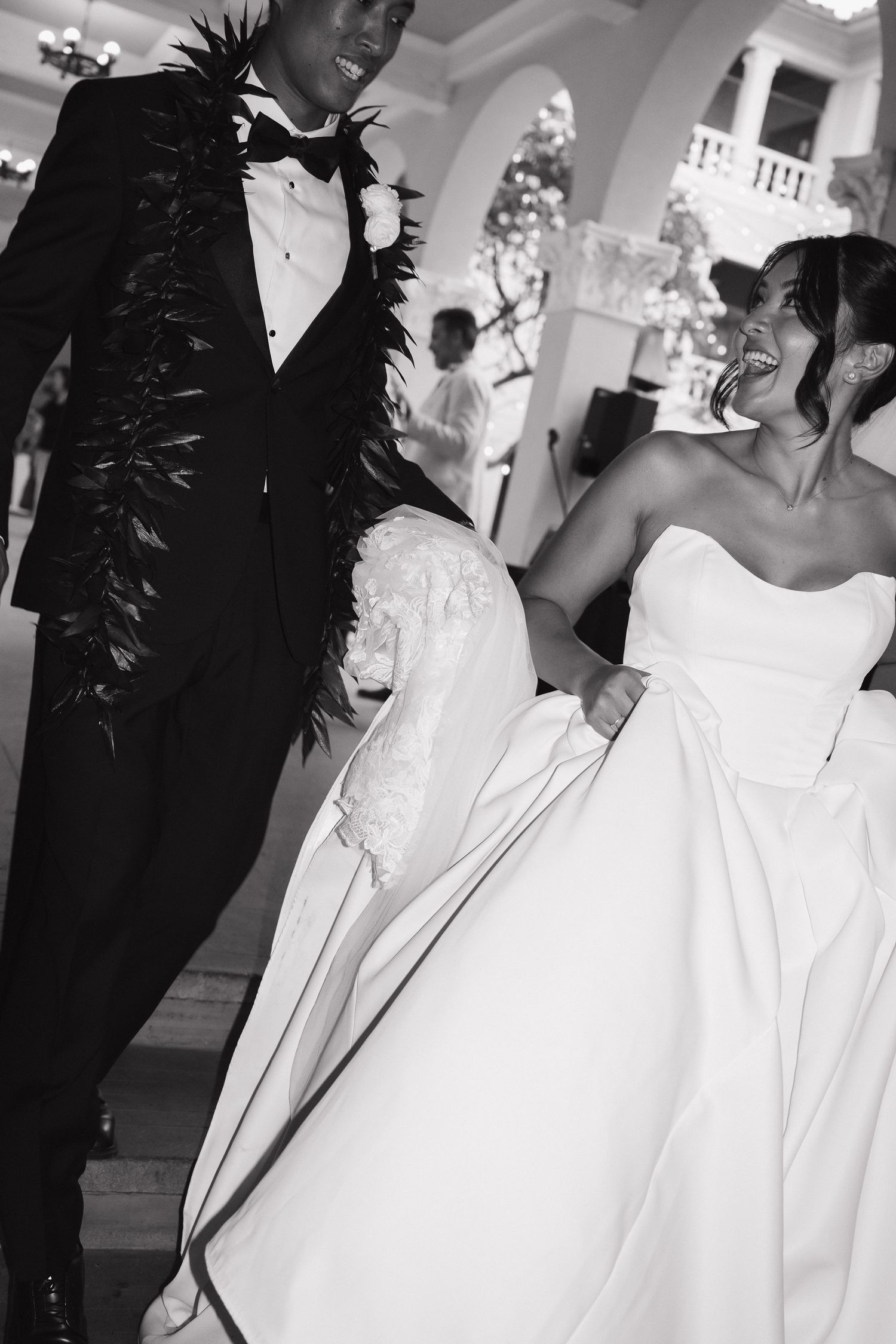 Black and white photo of a joyful bride in a strapless gown and groom in a tuxedo with floral lei at their wedding.