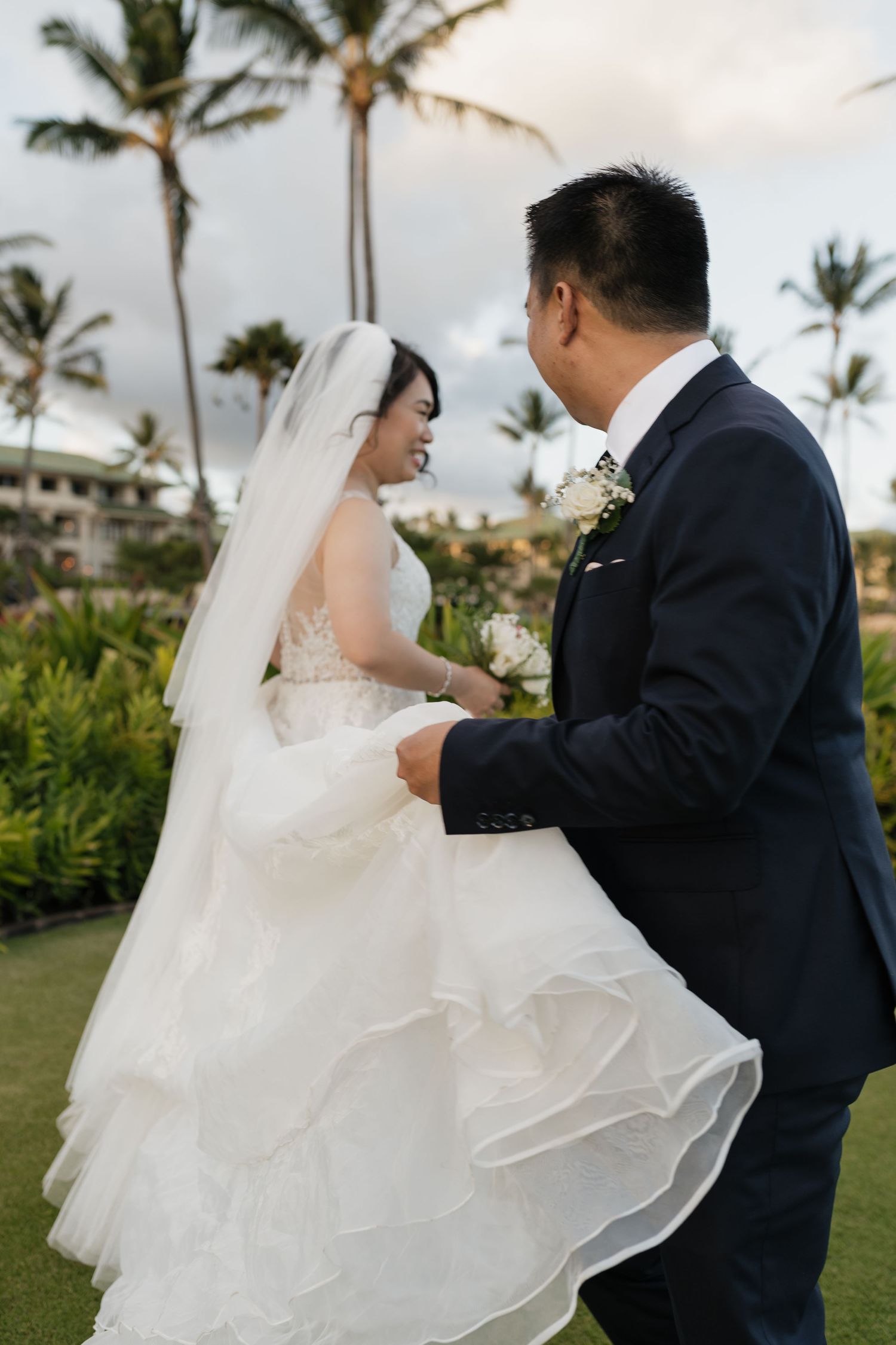 Newlyweds in formal attire sharing playful moments on a grassy lawn with palm trees.