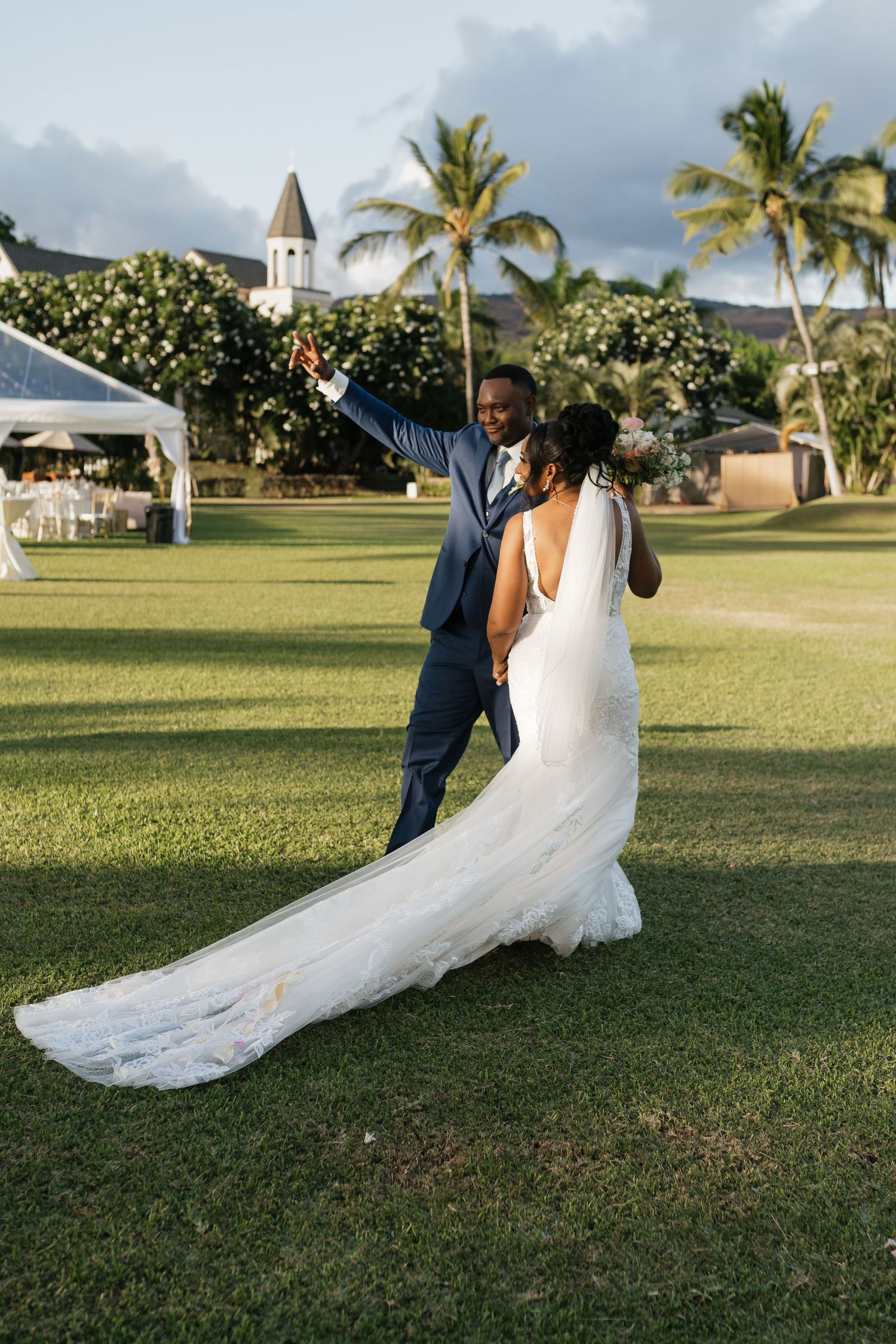Wedding couple walking across a lush green lawn with flowing white dress trailing behind.