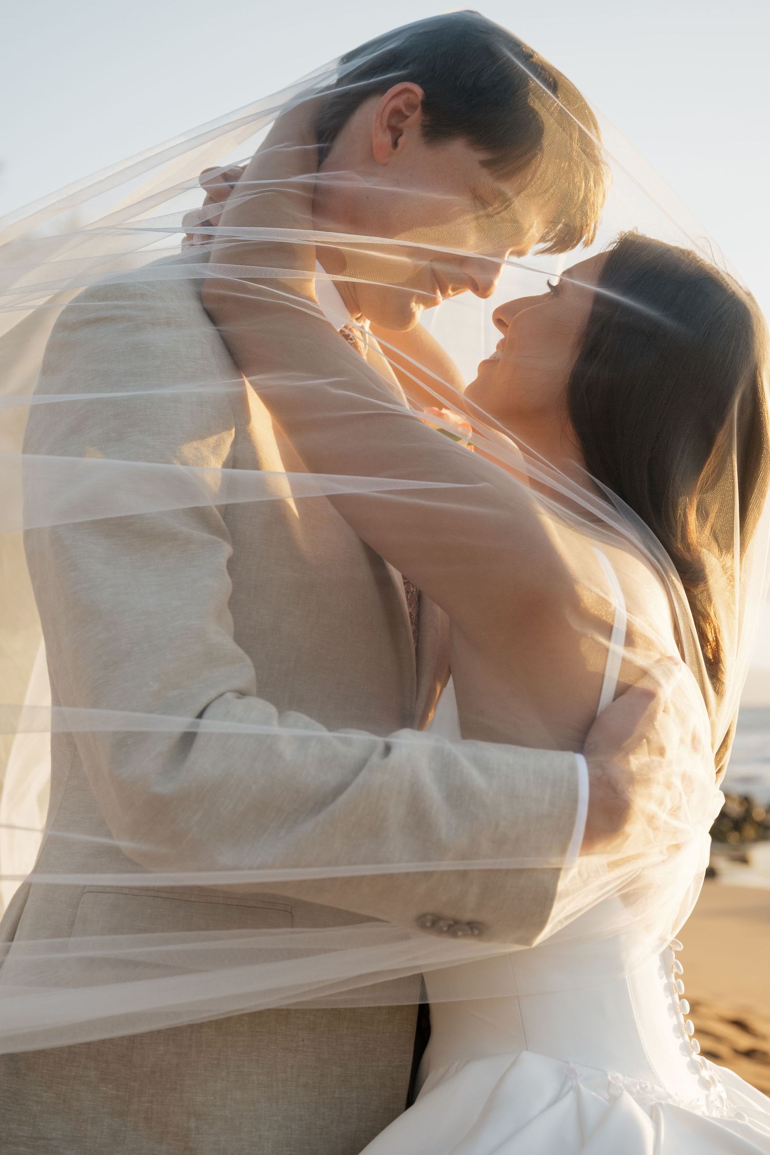 Romantic silhouette of a couple embraced under a flowing veil at sunset on the beach.