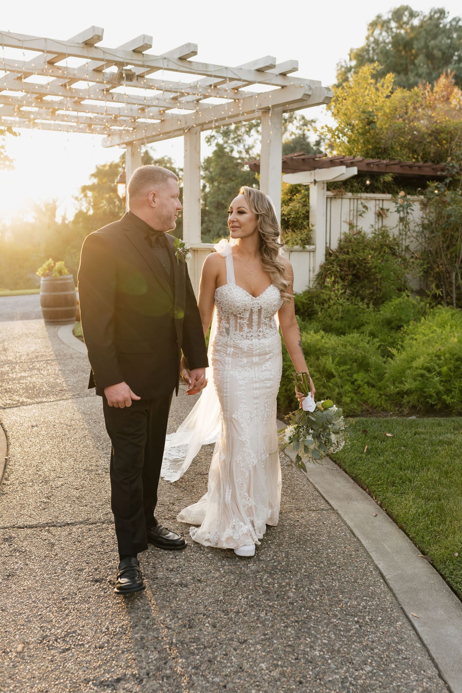 A couple walks hand in hand under a garden pergola at sunset in formal wedding attire.