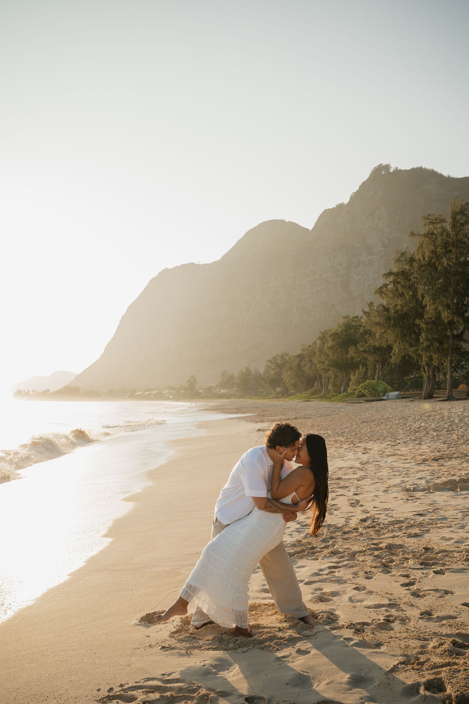 A couple embraces on a scenic beach at sunset with mountains and palm trees in the background.