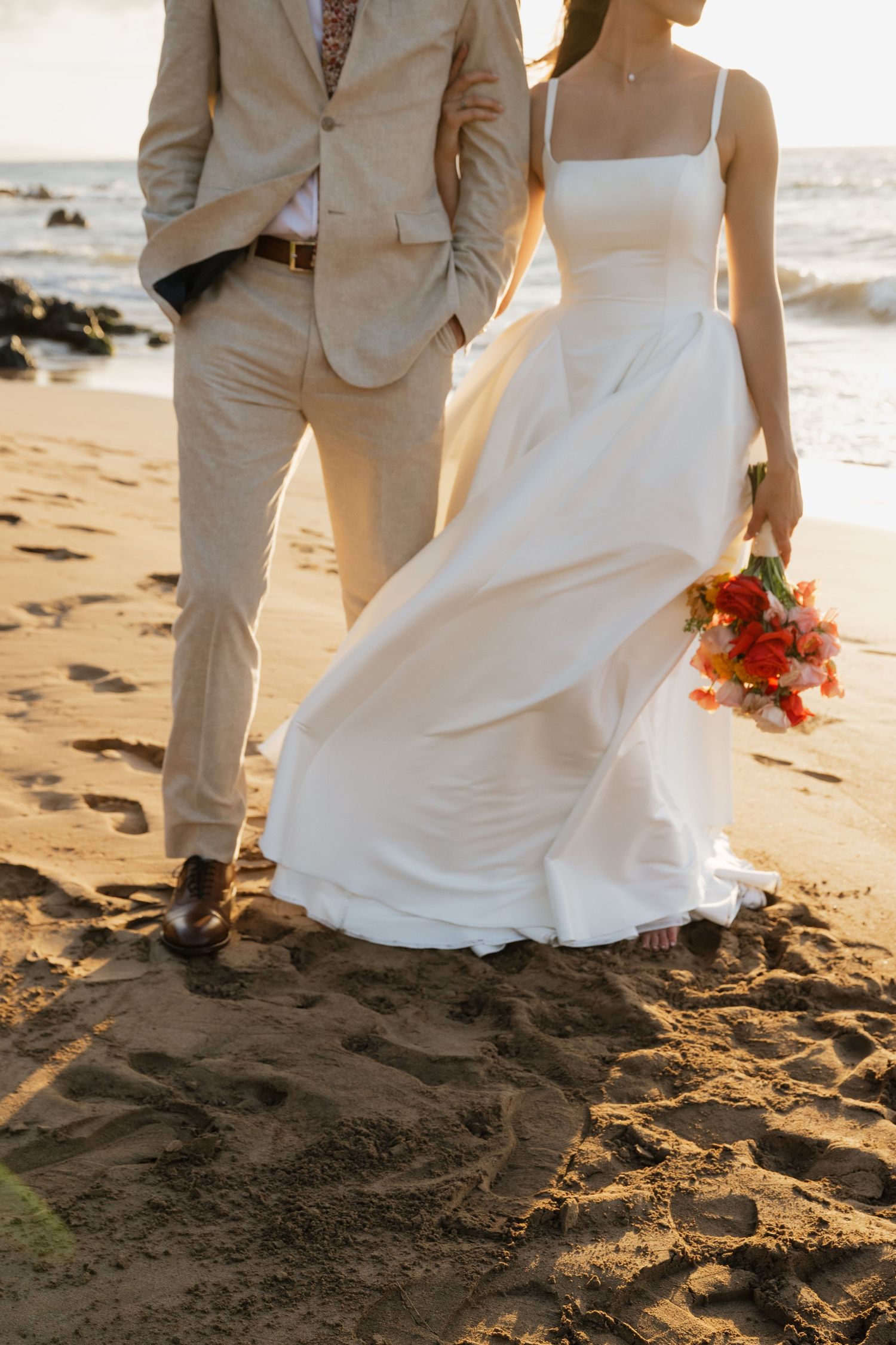 Couple walking on sandy beach with bride holding orange flowers at sunset.