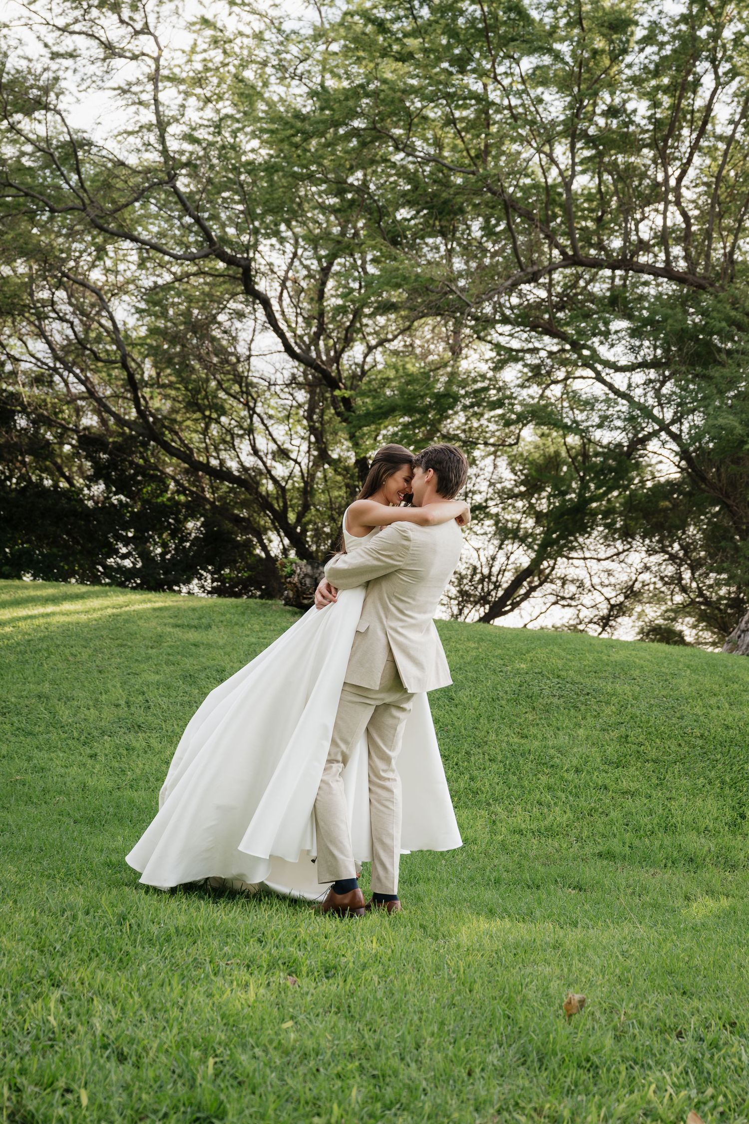 Two brides in white dresses twirl and dance together on a grassy hill during their wedding celebration.