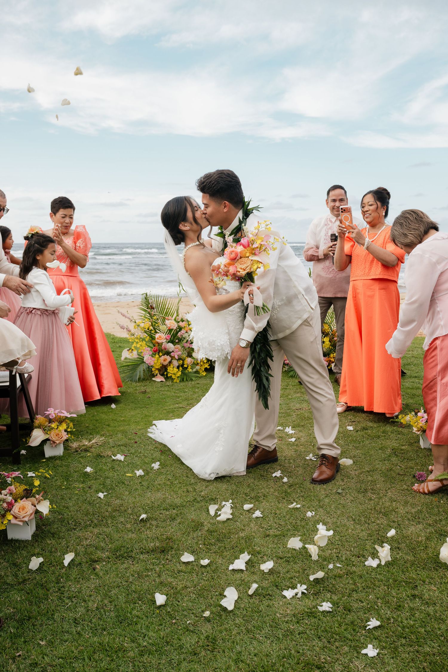 Newlyweds share first kiss on oceanfront lawn while guests in coral dresses celebrate with rose petals.