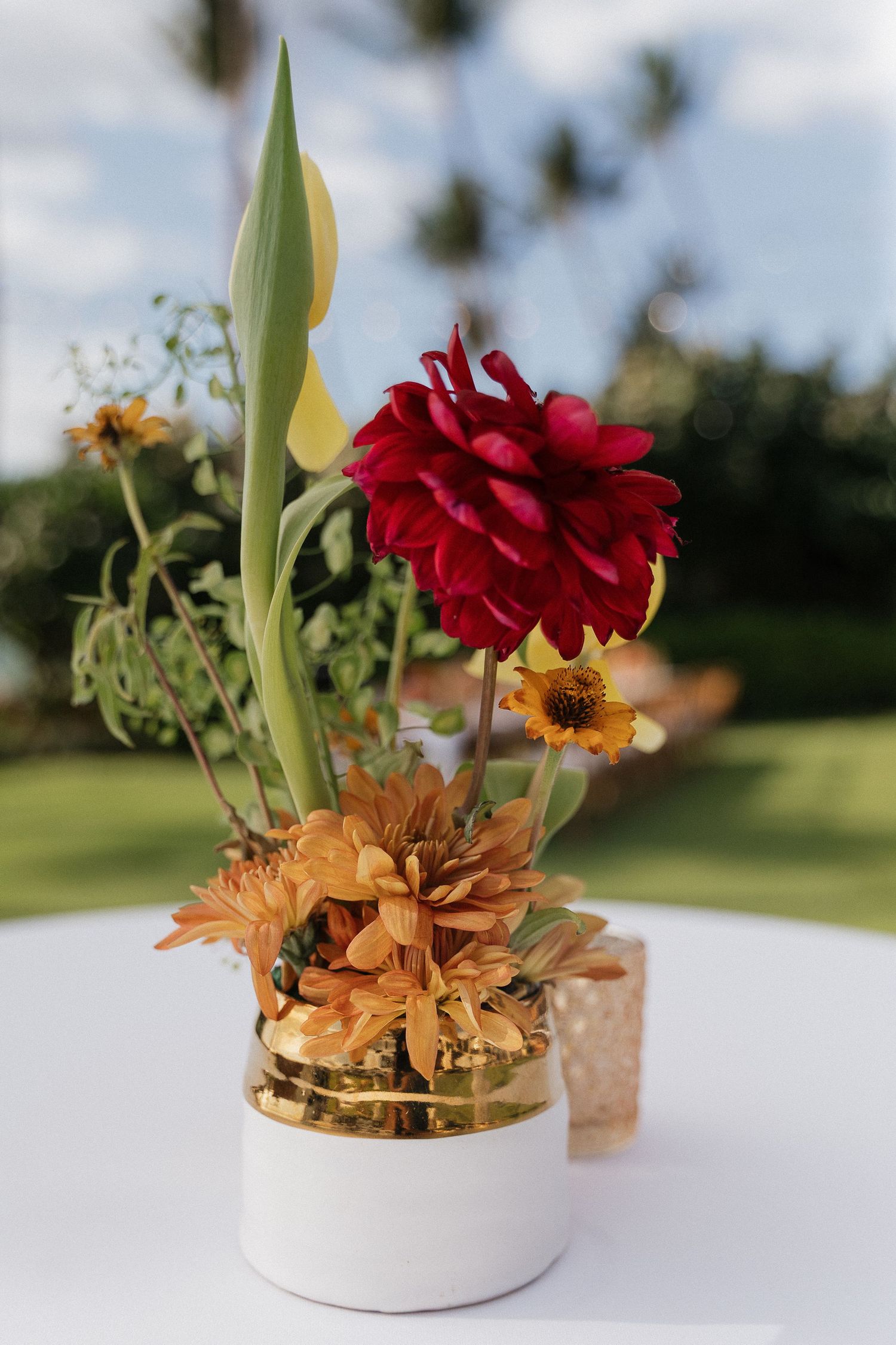 Colorful floral centerpiece arrangement with red and orange blooms in a white ceramic vessel on a light surface.