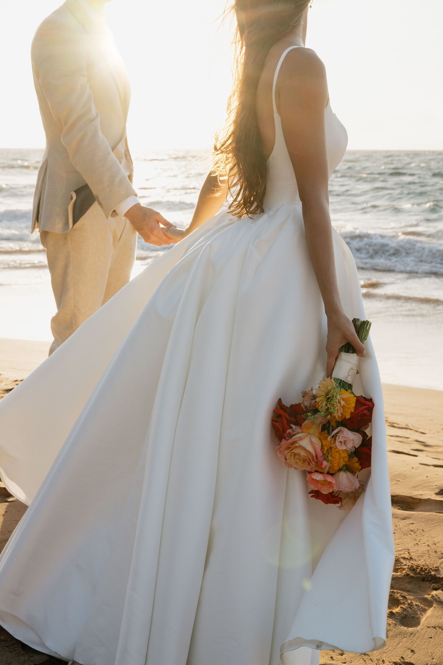 Close-up of a white wedding dress and autumn-colored bouquet against a beach sunset backdrop.