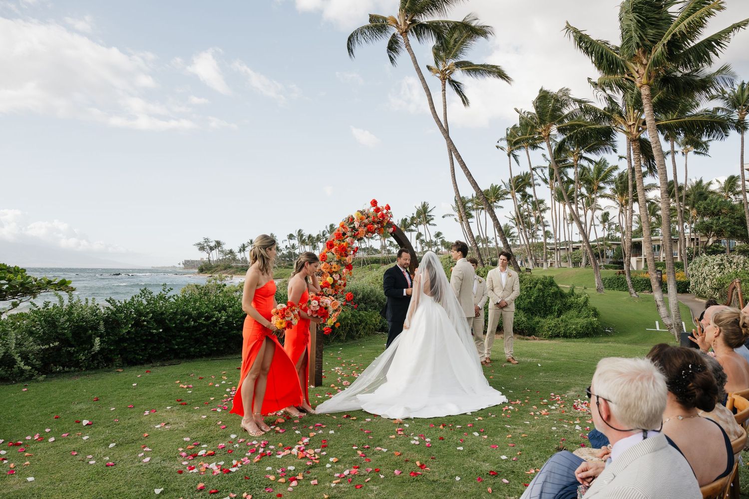 A tropical wedding ceremony on a grassy lawn with palm trees and flower petals scattered on the ground.