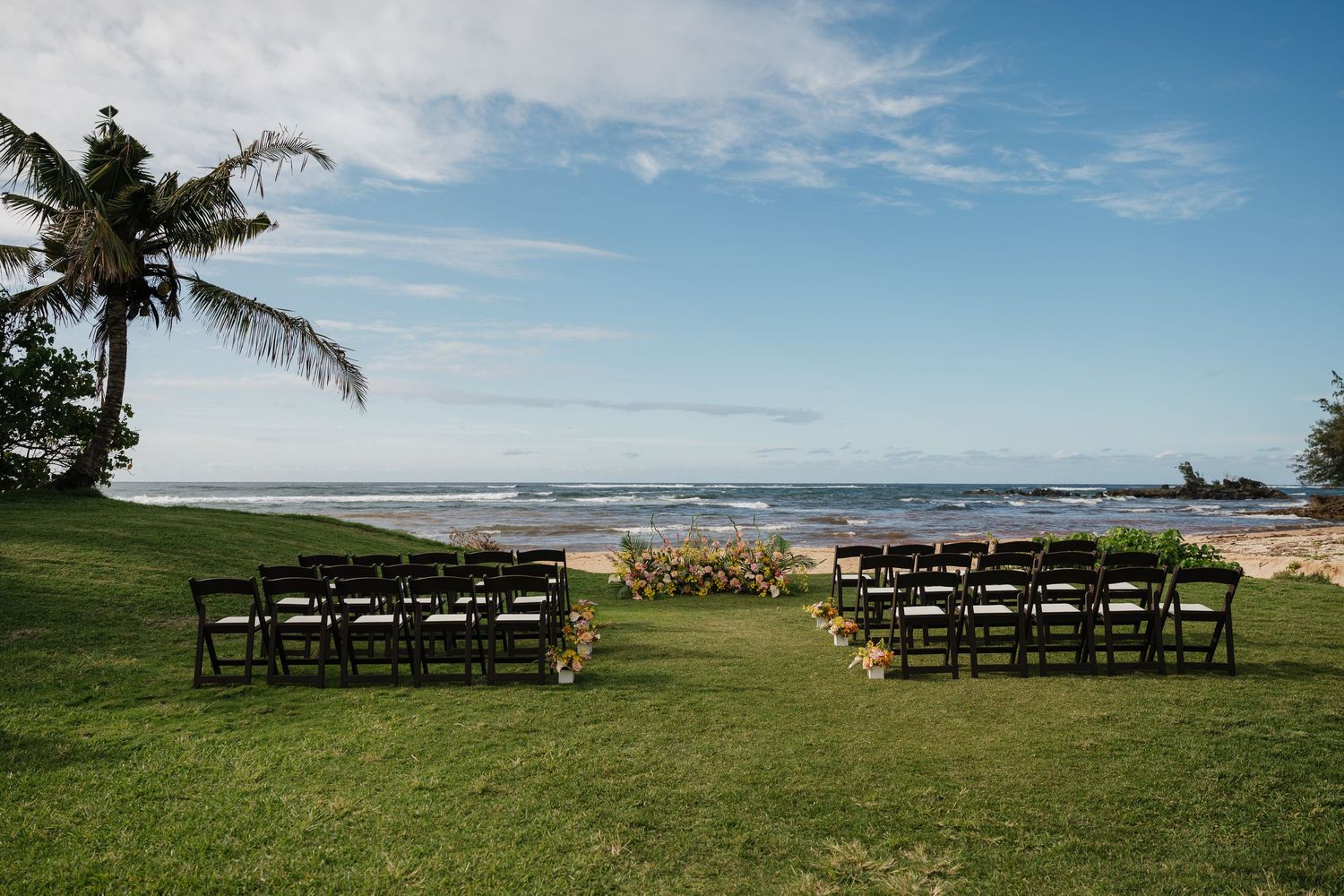 Outdoor beach wedding ceremony setup with dark chairs on green lawn, ocean backdrop, and floral arrangements under blue sky.