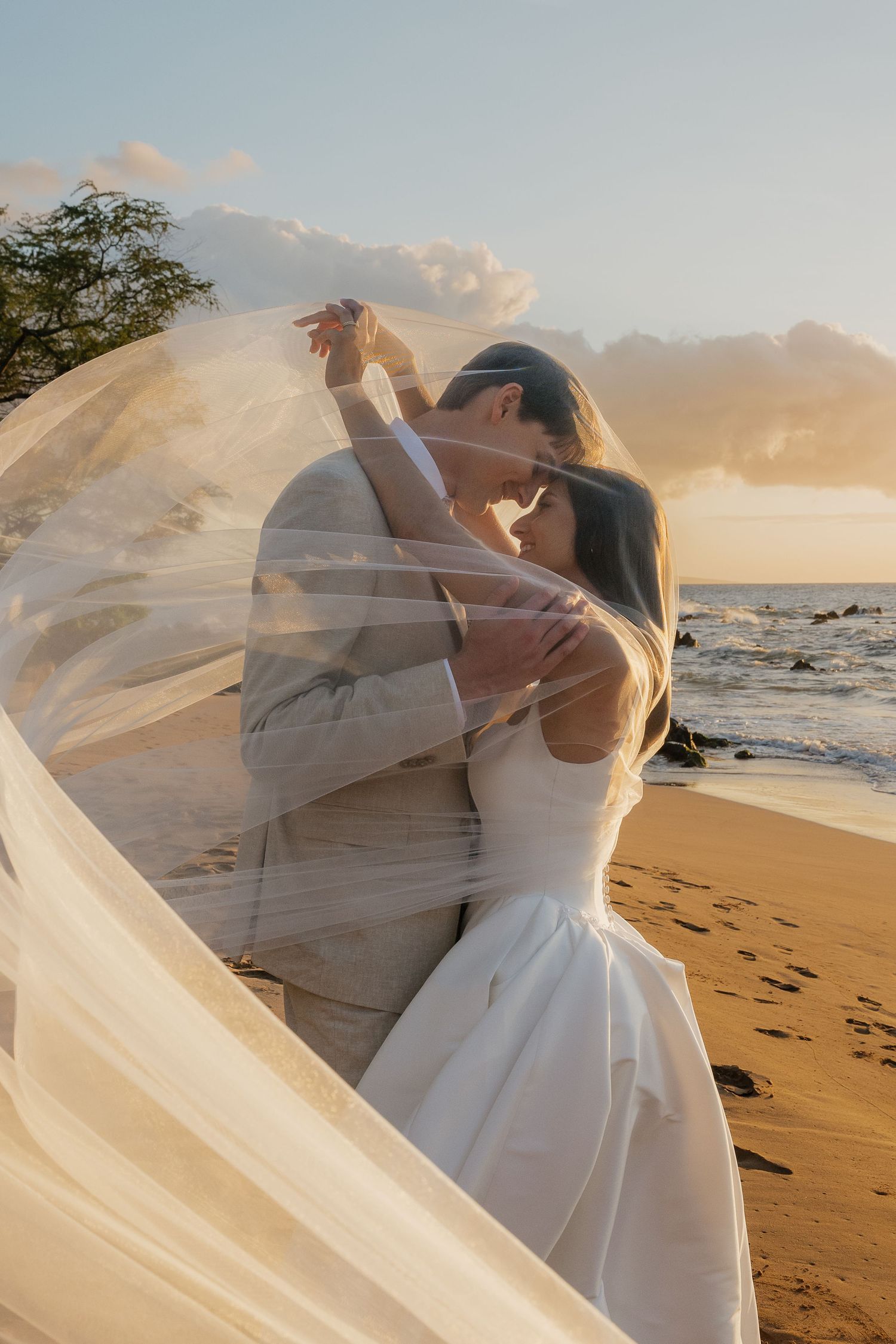 A romantic beach wedding photoshoot captures a couple embracing as a flowing white veil catches the golden sunset light.