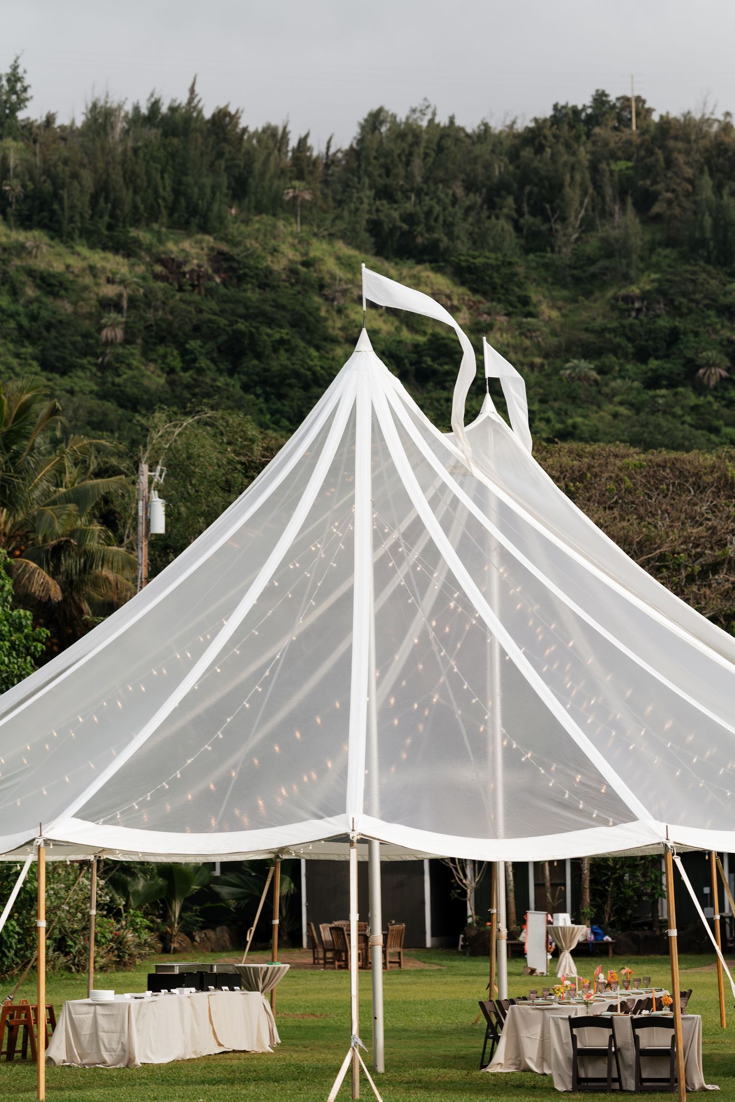 White sailcloth wedding tent with string lights on lush green lawn with tropical hillside backdrop.