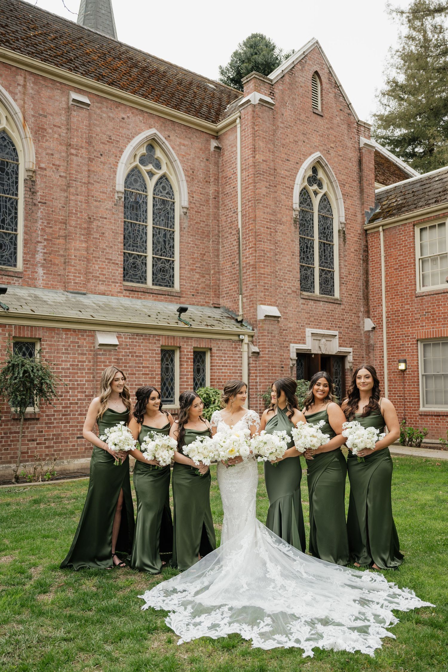 Wedding party in emerald green dresses poses outside a brick church building.