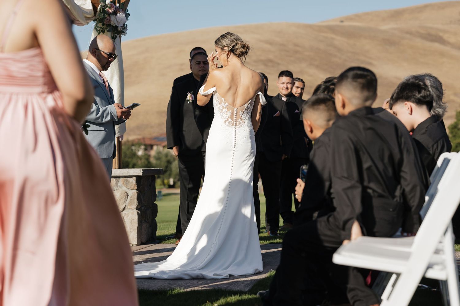 Wedding ceremony attendees gather outdoors in formal attire against scenic rolling hills.
