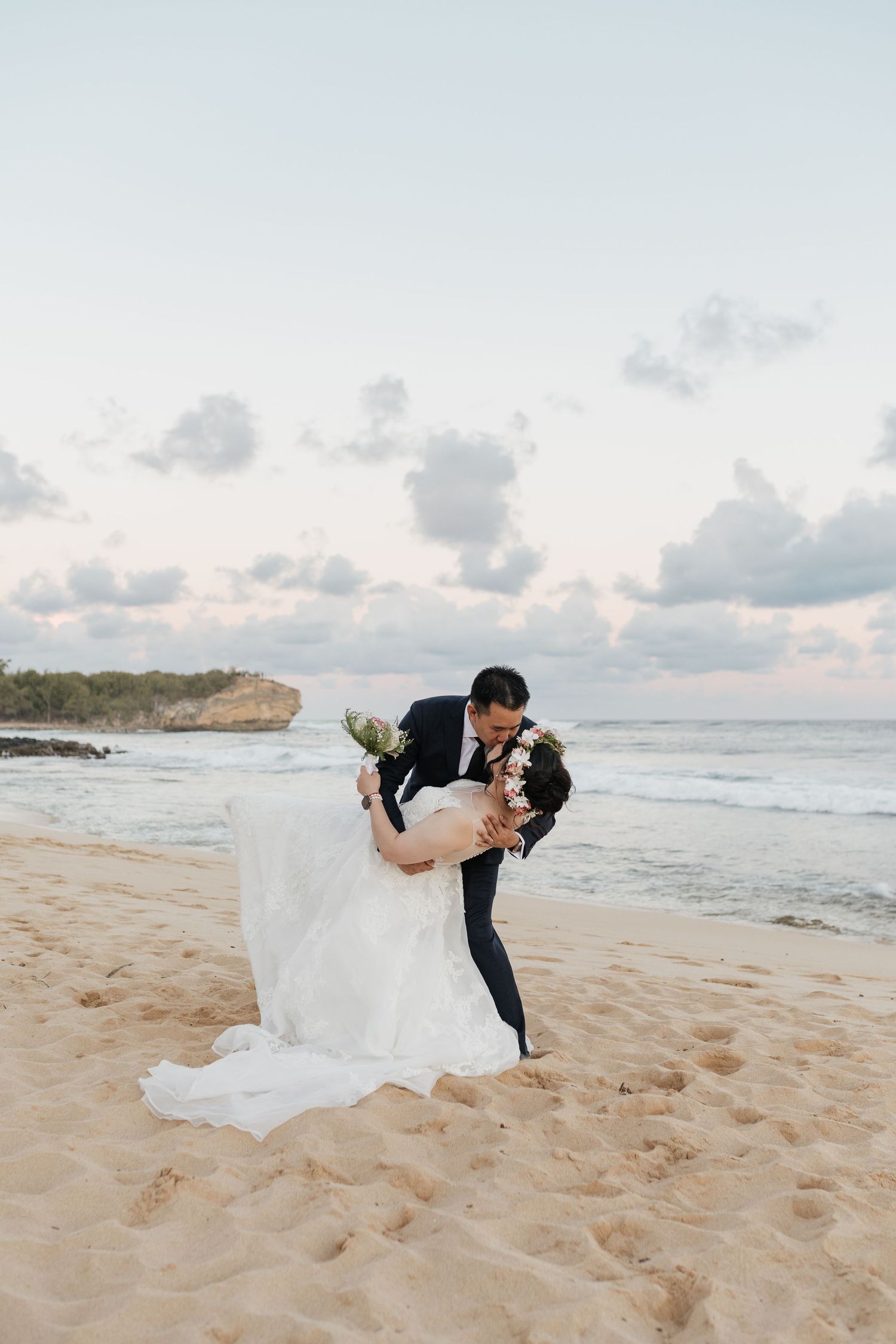 A romantic beach wedding dip pose with waves and cliffs in background.