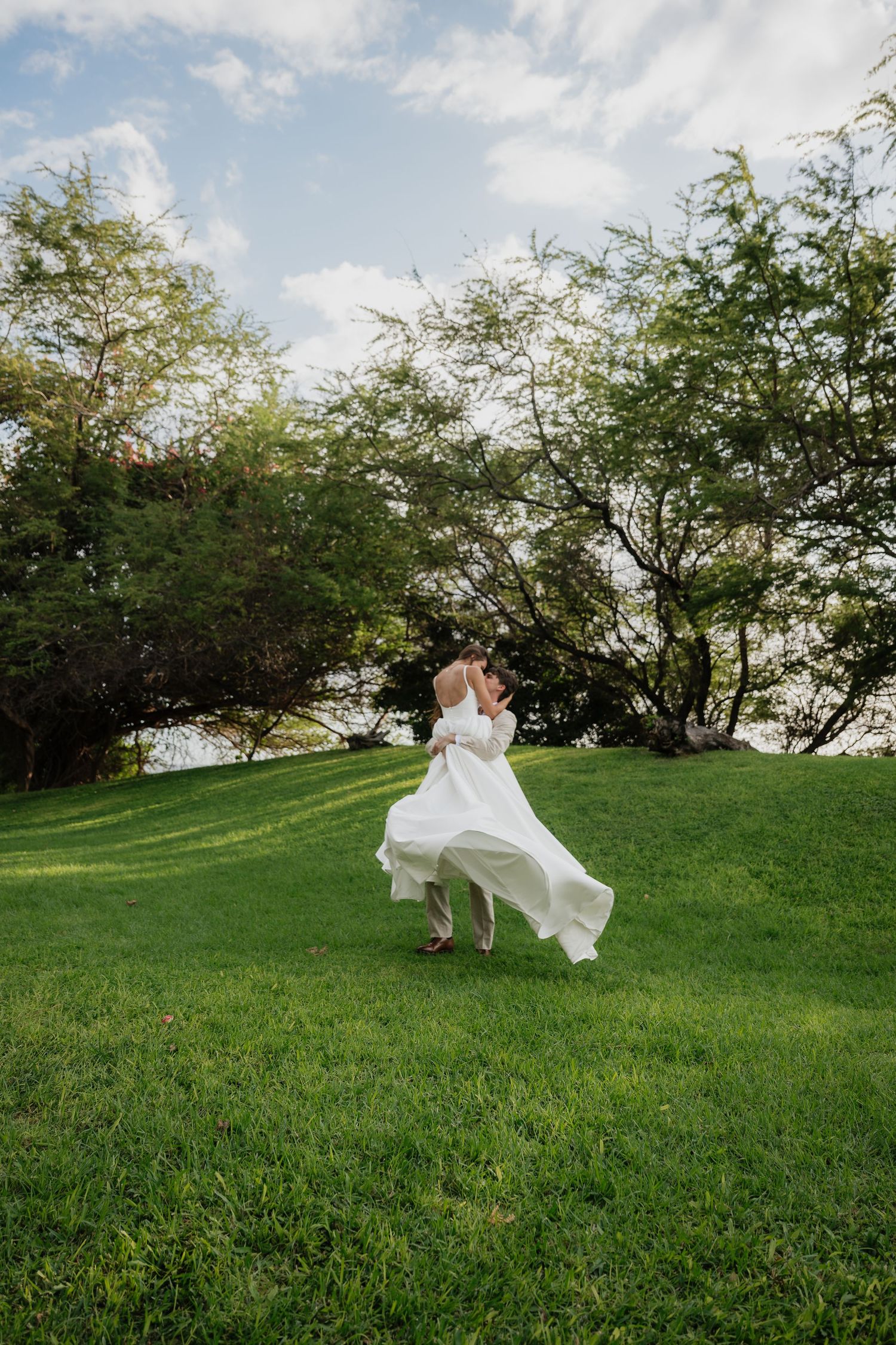 Wedding dress flows dramatically across green hillside under blue sky.