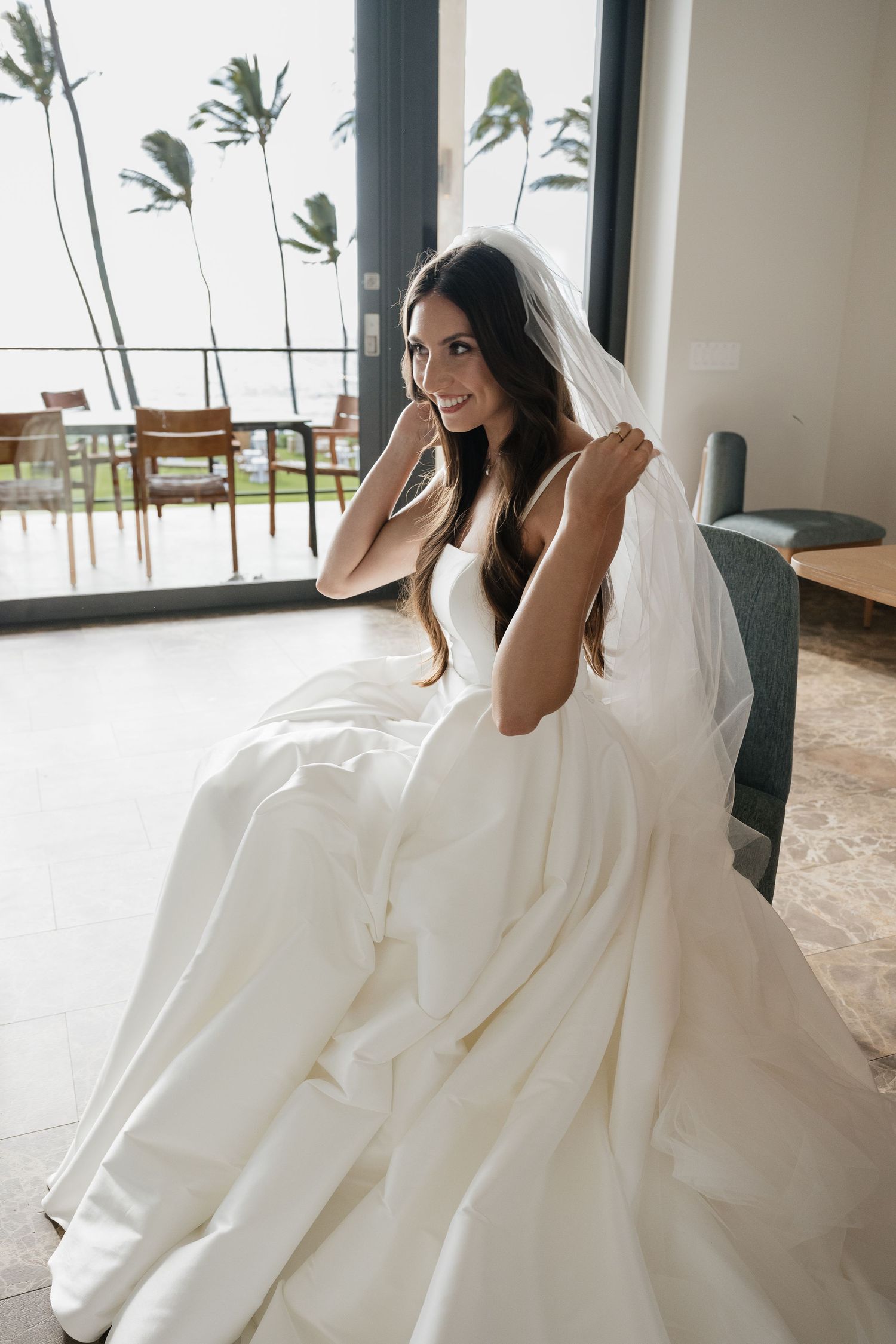 A bride in a flowing white gown poses playfully on a couch near large windows with palm trees visible outside.